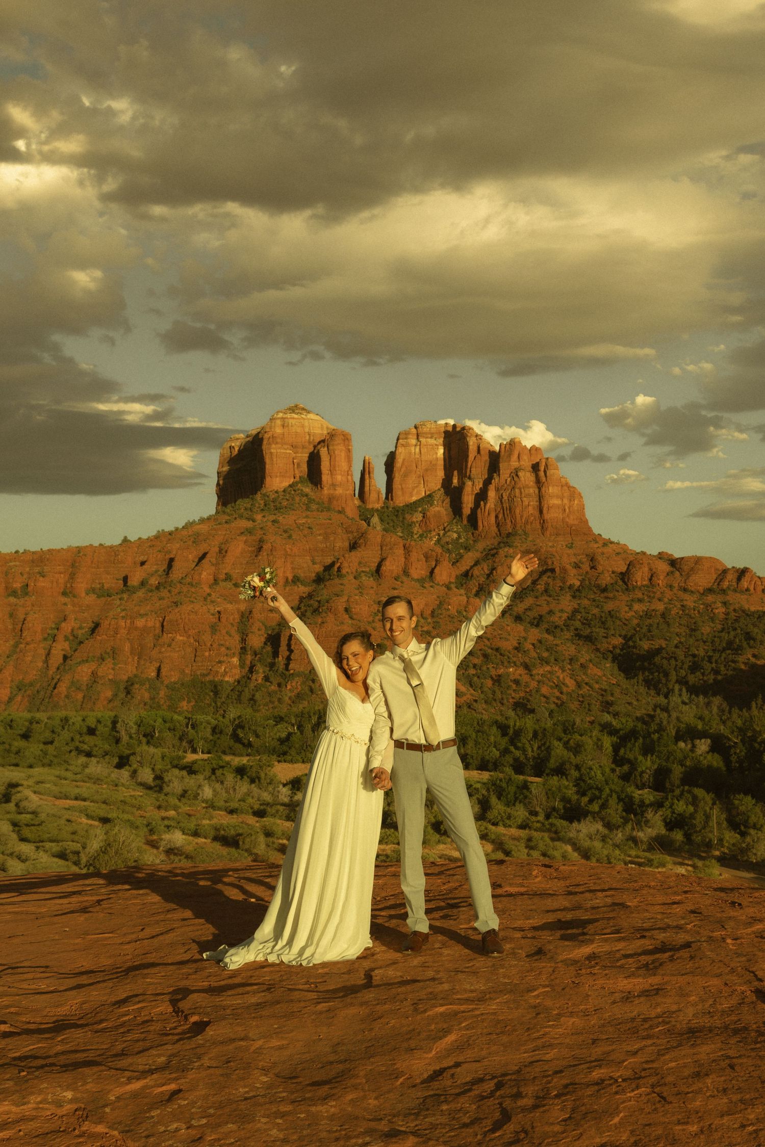 Joyful couple in wedding attire celebrating at Cathedral Rock in Sedona, Arizona, with red rock formations at sunset.