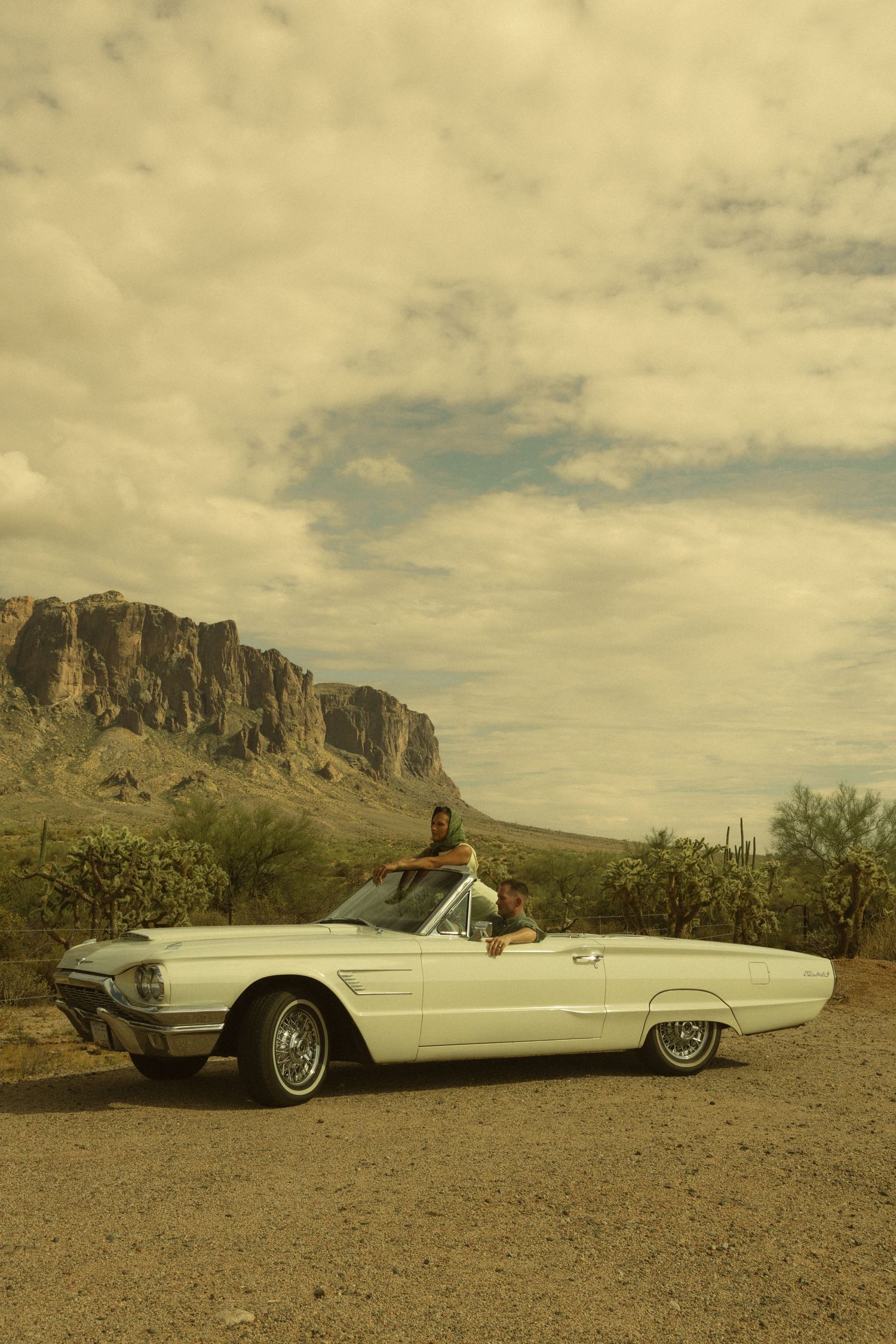 A woman poses in a white vintage Ford Thunderbird convertible on a desert road with rocky mountains in the background.