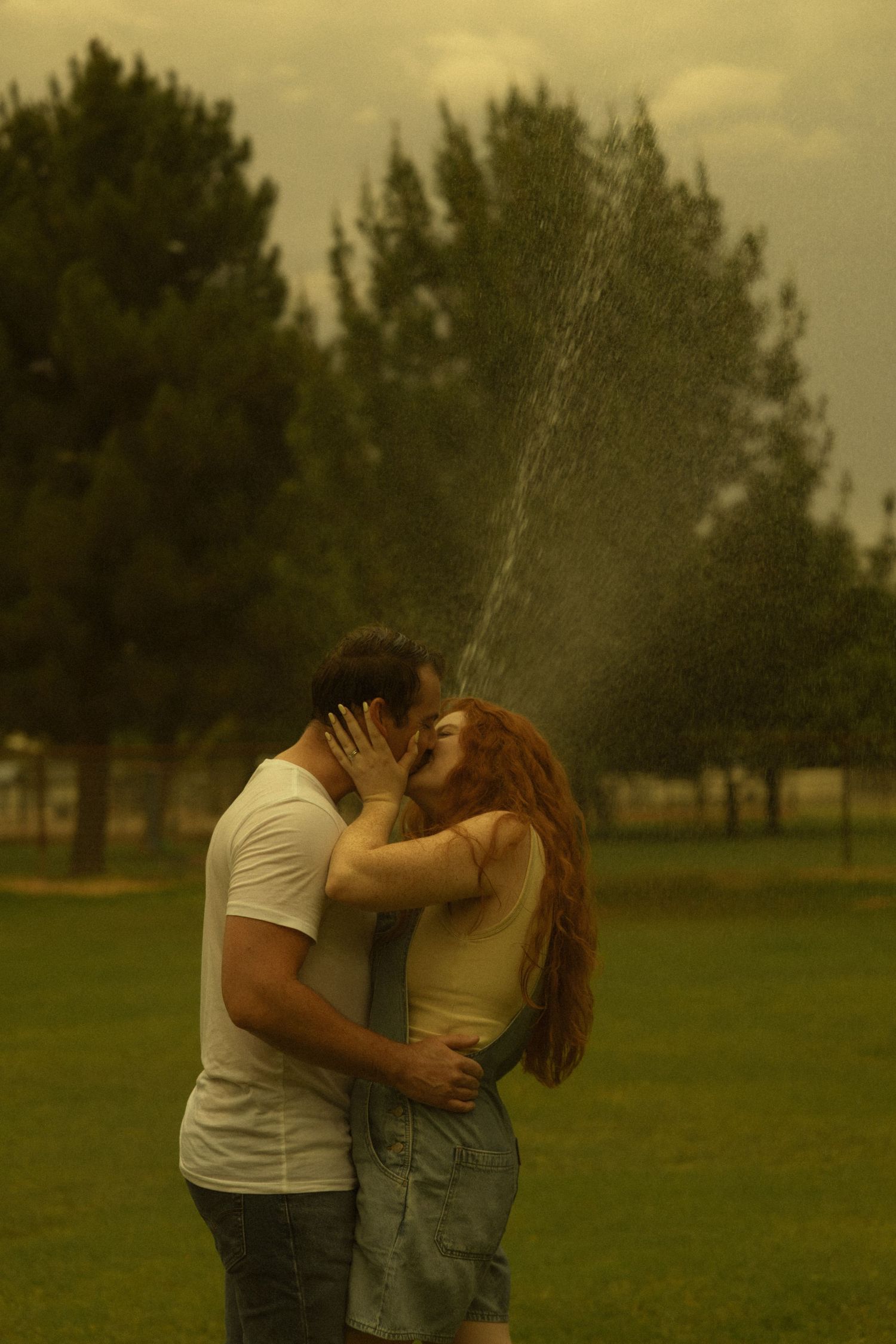 Couple kissing romantically in a park while water sprays around them under a dramatic golden sky.