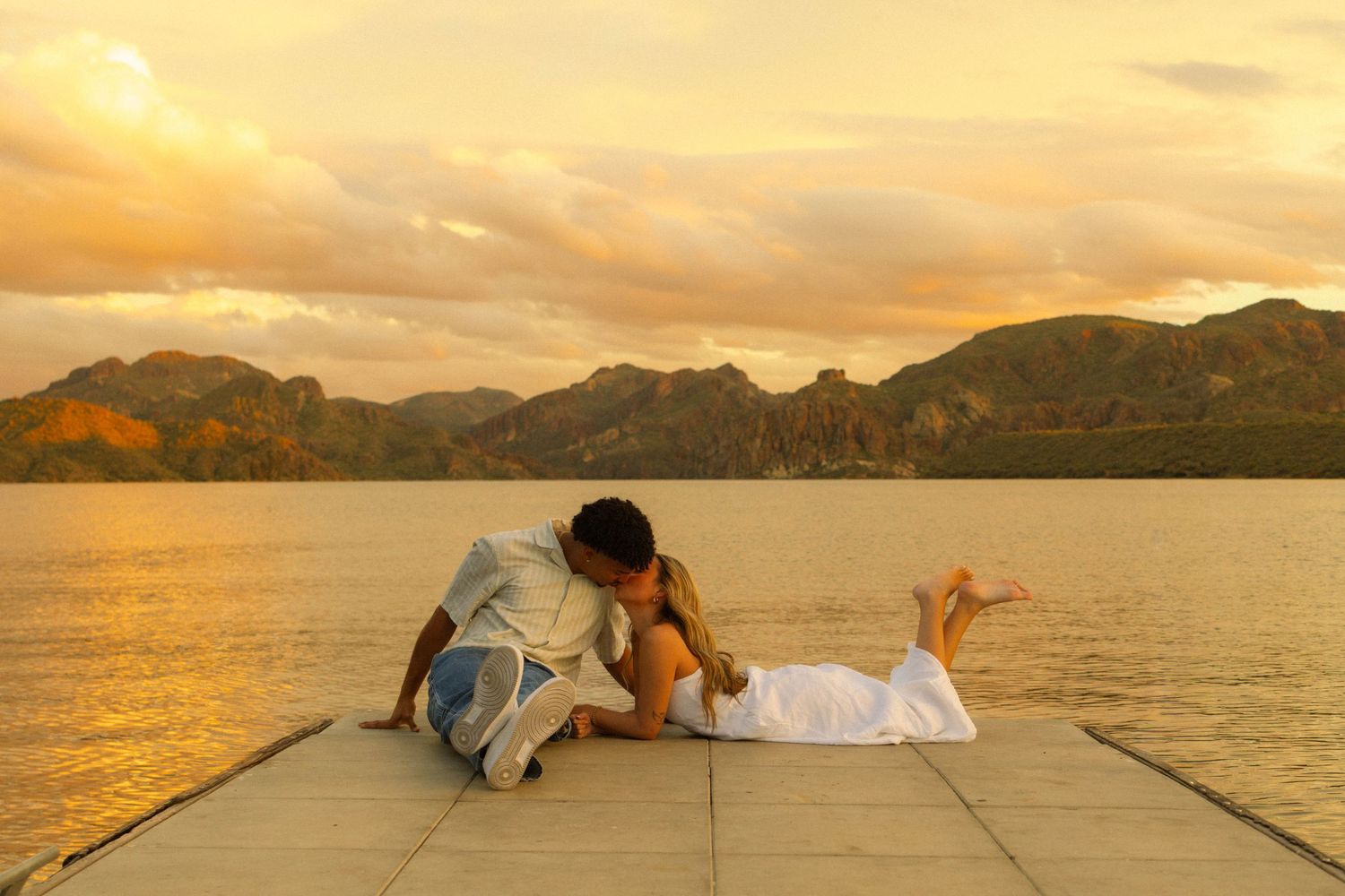Romantic couple embraces on a pier at sunset with mountains and ocean in the background creating a dreamy scene.