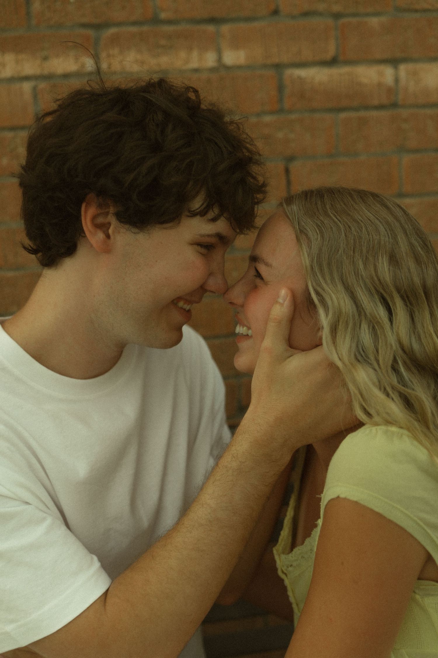 Young couple smiling and touching foreheads tenderly against a brick wall in a romantic close-up.