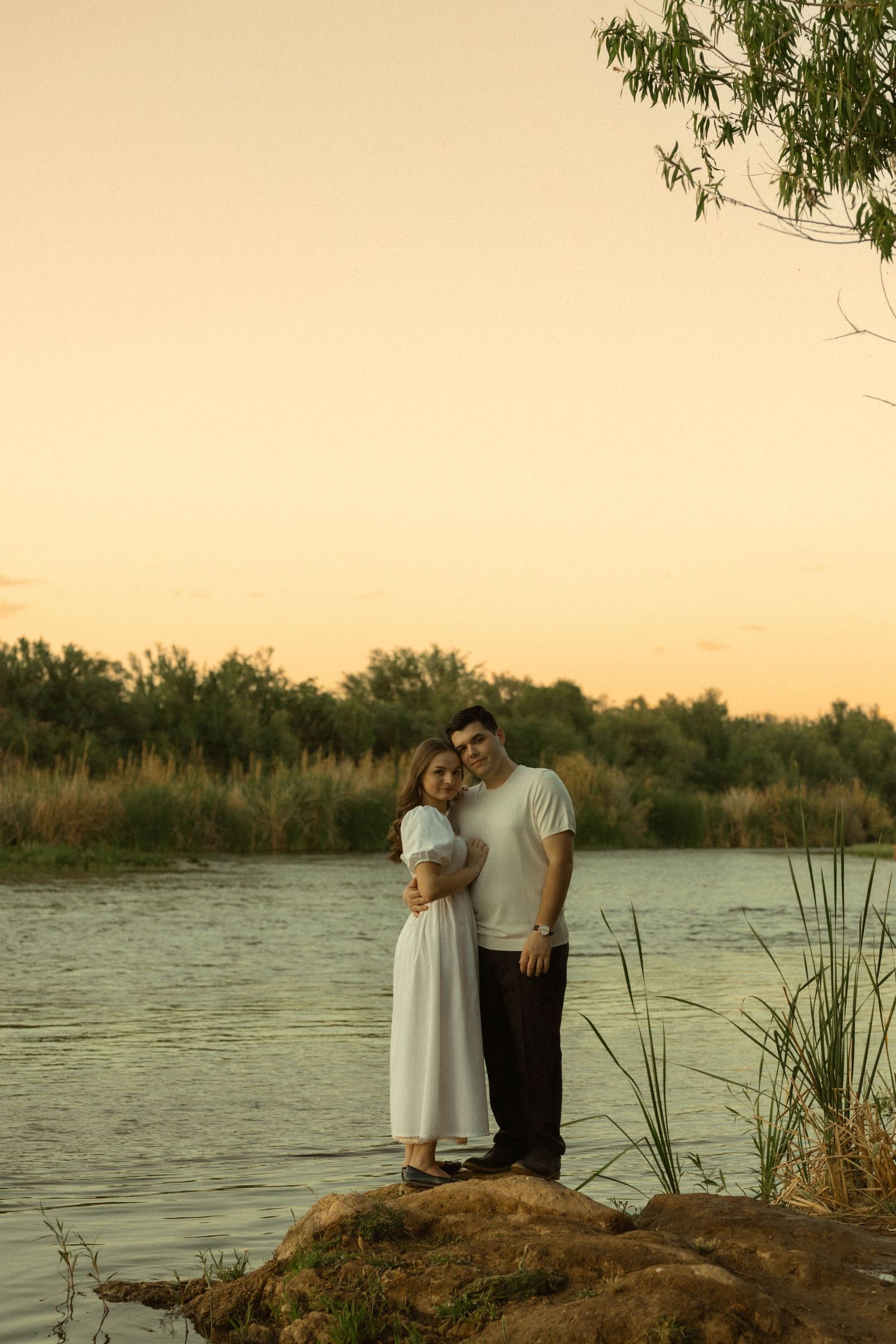 Happy couple embracing on a riverside rock at sunset, posing together in white dress and casual attire.