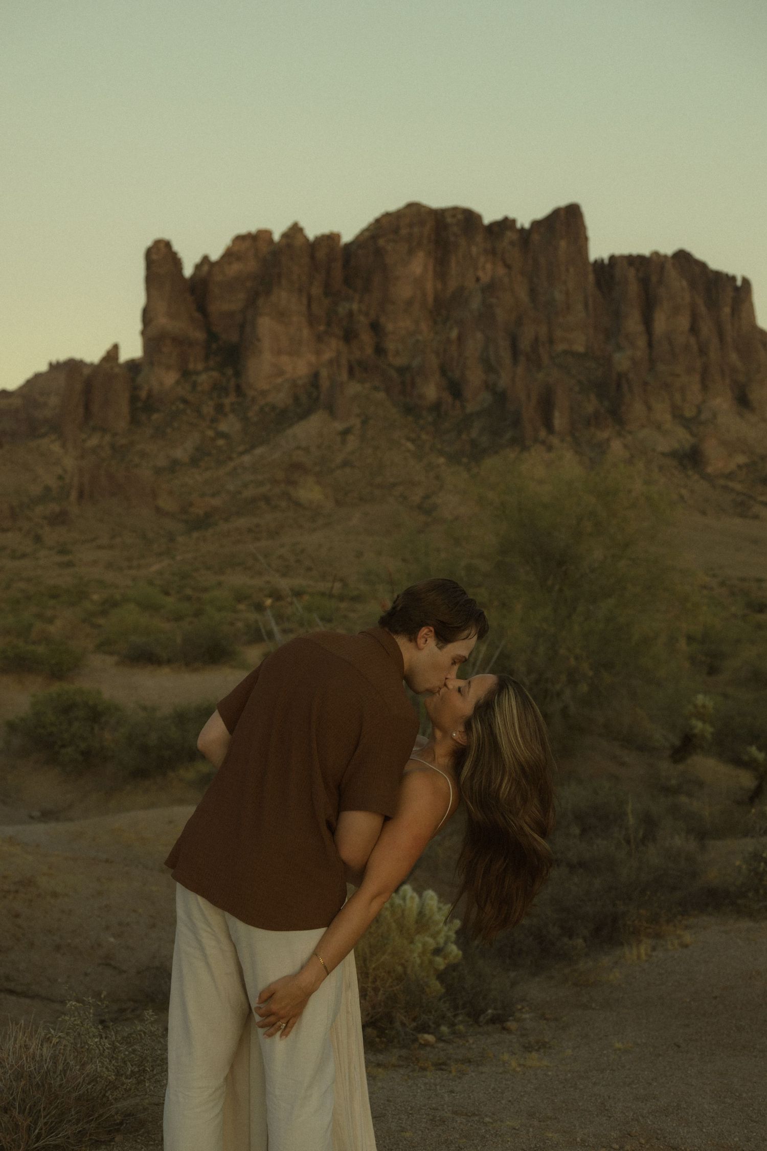 Man in brown shirt dipping partner for a kiss in desert landscape with rocky mountain backdrop at dusk.