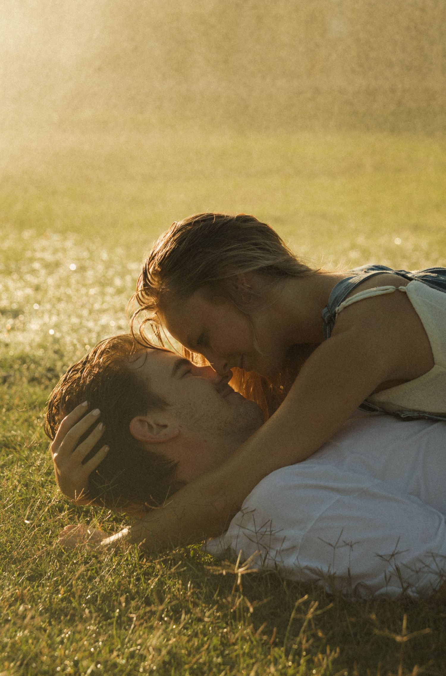 A couple shares an intimate kiss lying on the grass bathed in warm golden sunlight.
