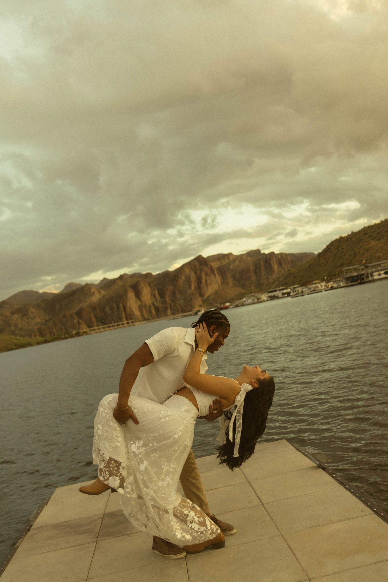 Couple sharing a romantic dip kiss on a waterfront dock with mountain lake backdrop under dramatic cloudy sky.