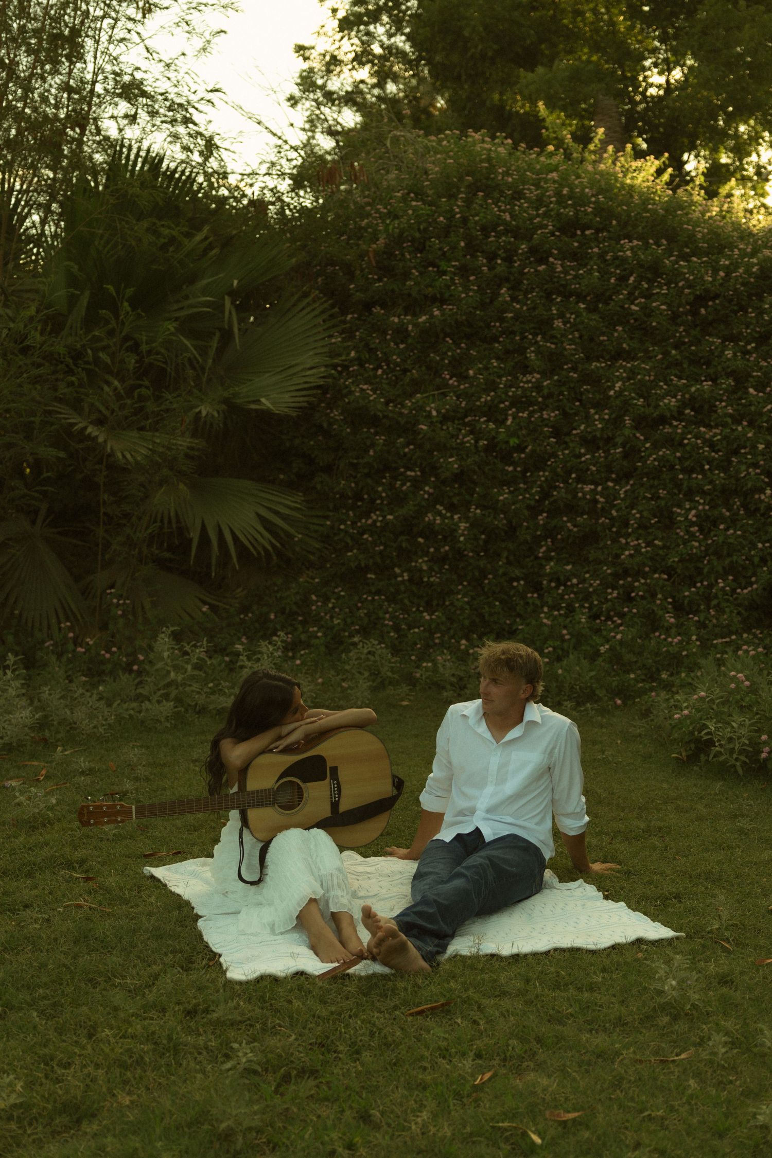 Couple relaxing on a white blanket in a lush garden, with an acoustic guitar leaning against the woman in a white dress.
