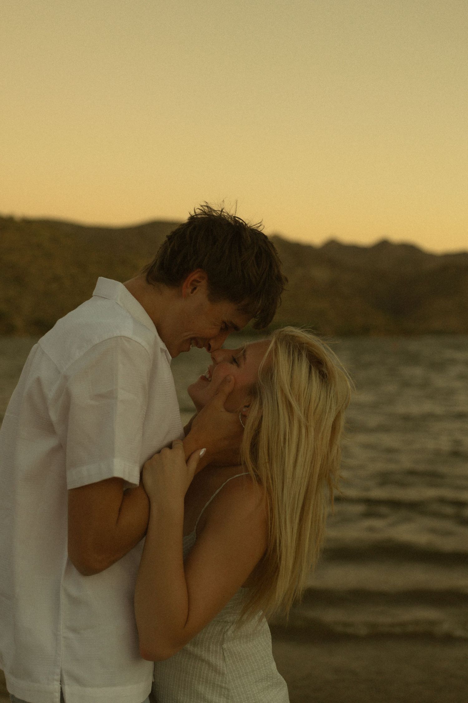 Romantic couple sharing an intimate moment by a lake at golden sunset, woman with blonde hair, man in white shirt.