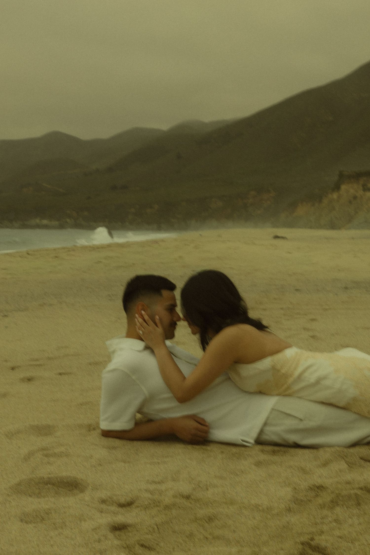 Couple lying on sandy beach sharing a romantic moment with misty mountains and ocean waves in the background.