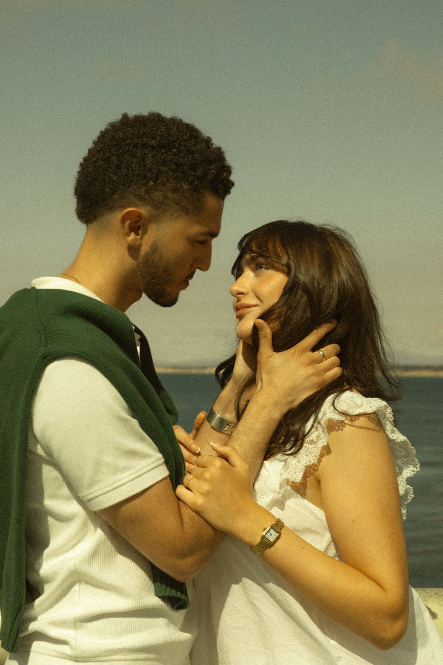 A romantic couple gazes at each other near the water, the woman in white lace and the man in a white tee with green sweater.