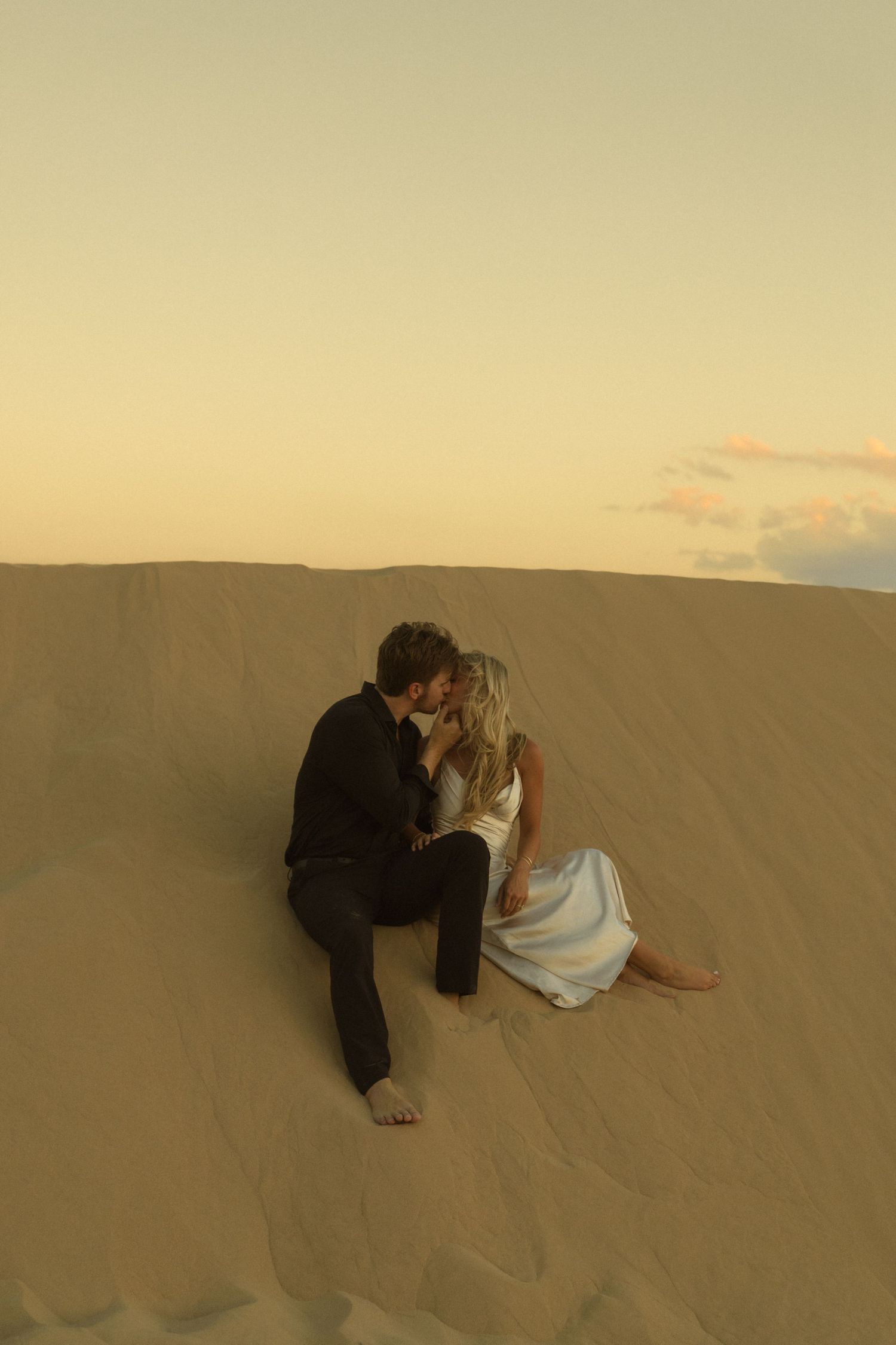 Couple sharing a romantic kiss while sitting on golden sand dunes at sunset in a desert landscape.