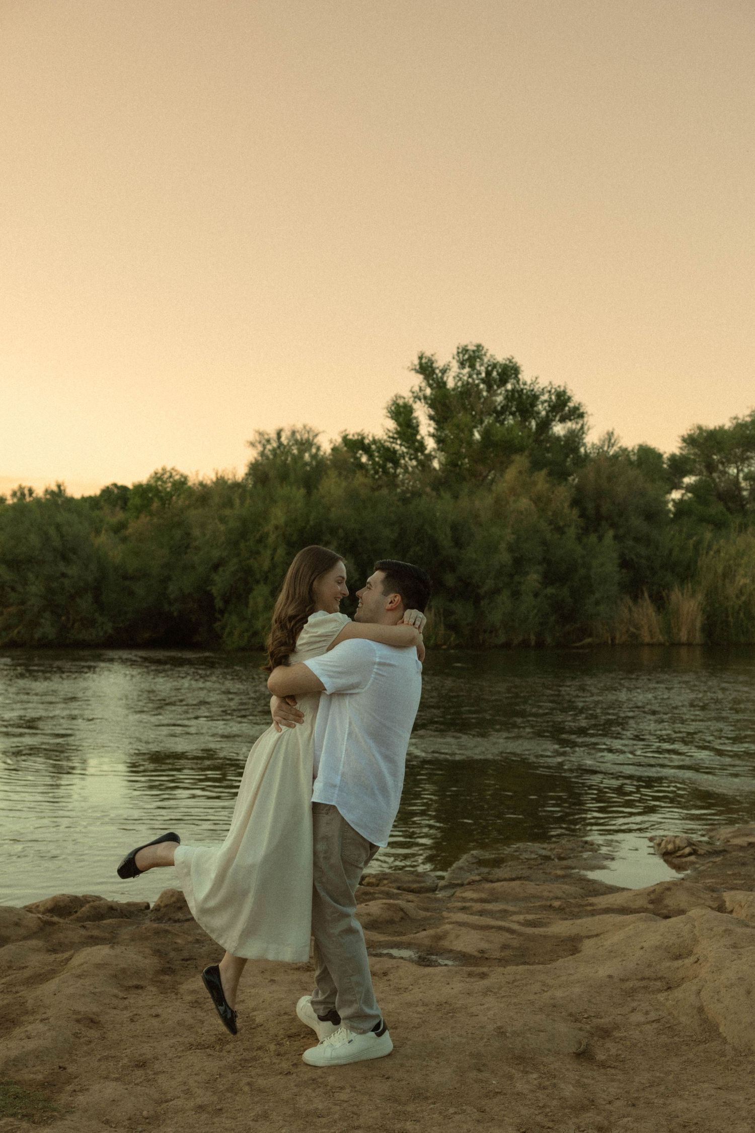 Couple embracing at sunset by a river, man lifting woman in white dress on sandy shore with green trees.
