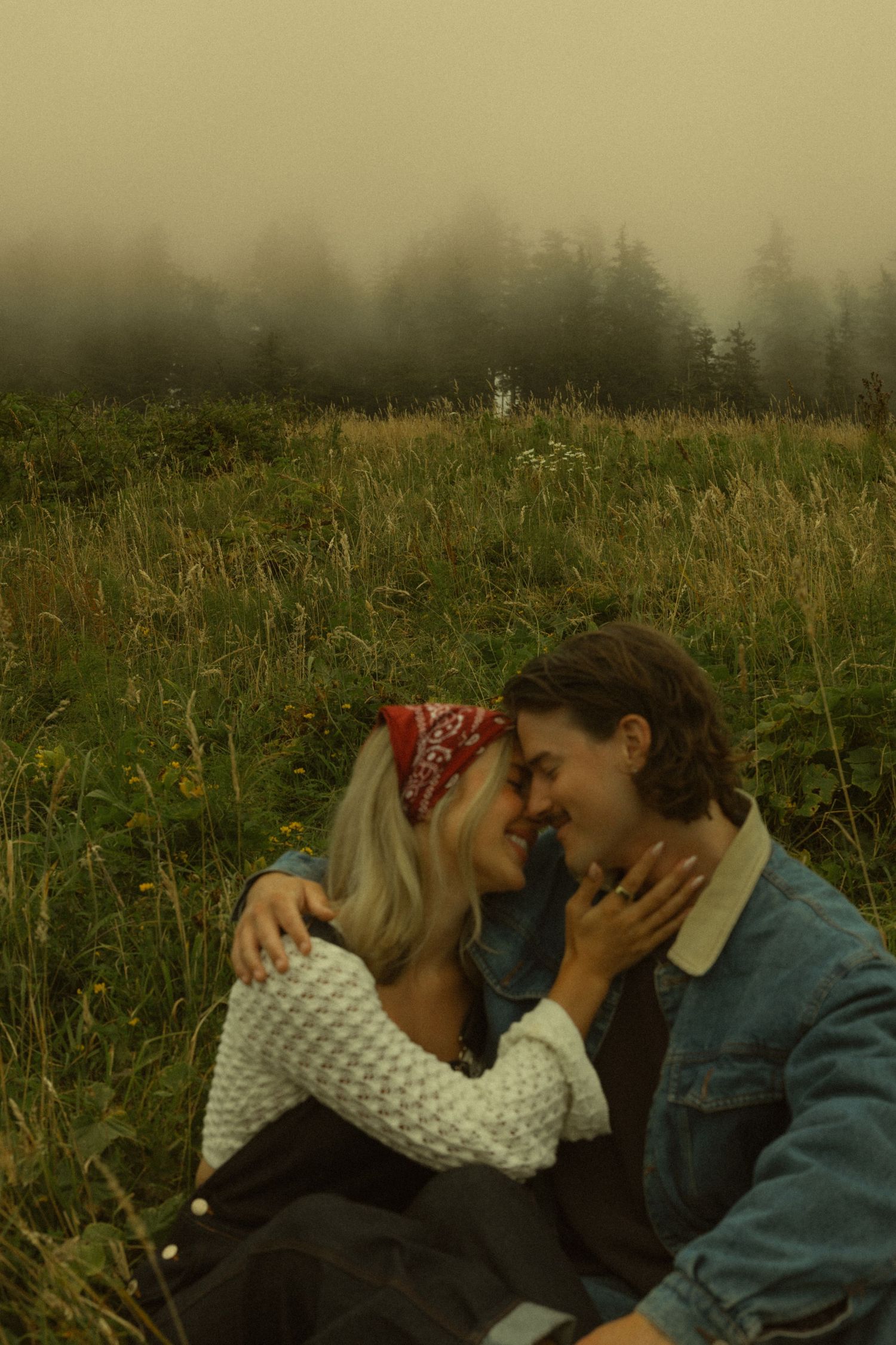 Couple sharing a romantic moment in a misty meadow, woman in red bandana and white sweater embracing man in denim jacket.