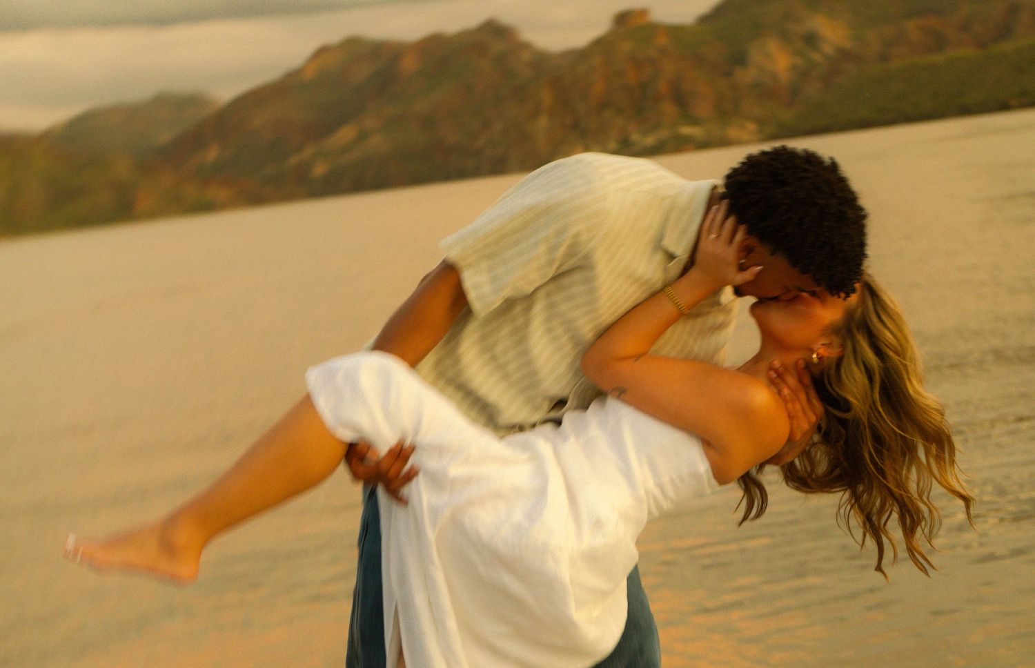 Romantic couple sharing a passionate kiss on sandy desert dunes with mountain backdrop at golden hour.