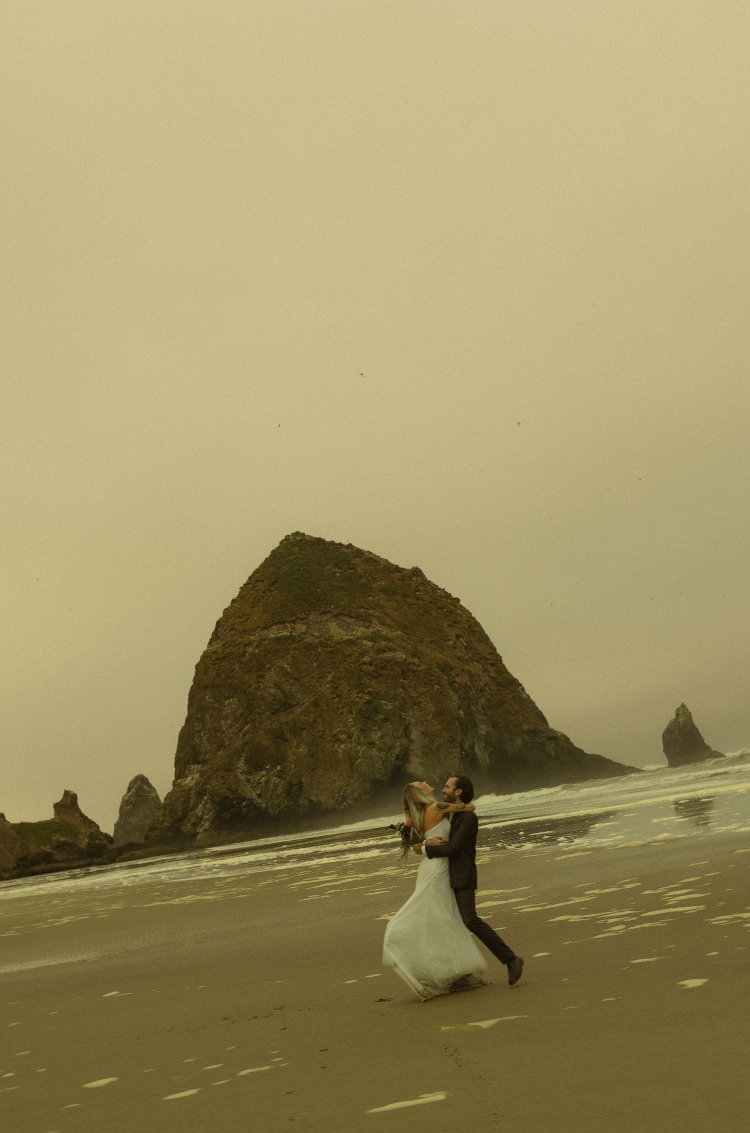 A couple in wedding attire dances romantically on a misty beach with Haystack Rock at Cannon Beach, Oregon.