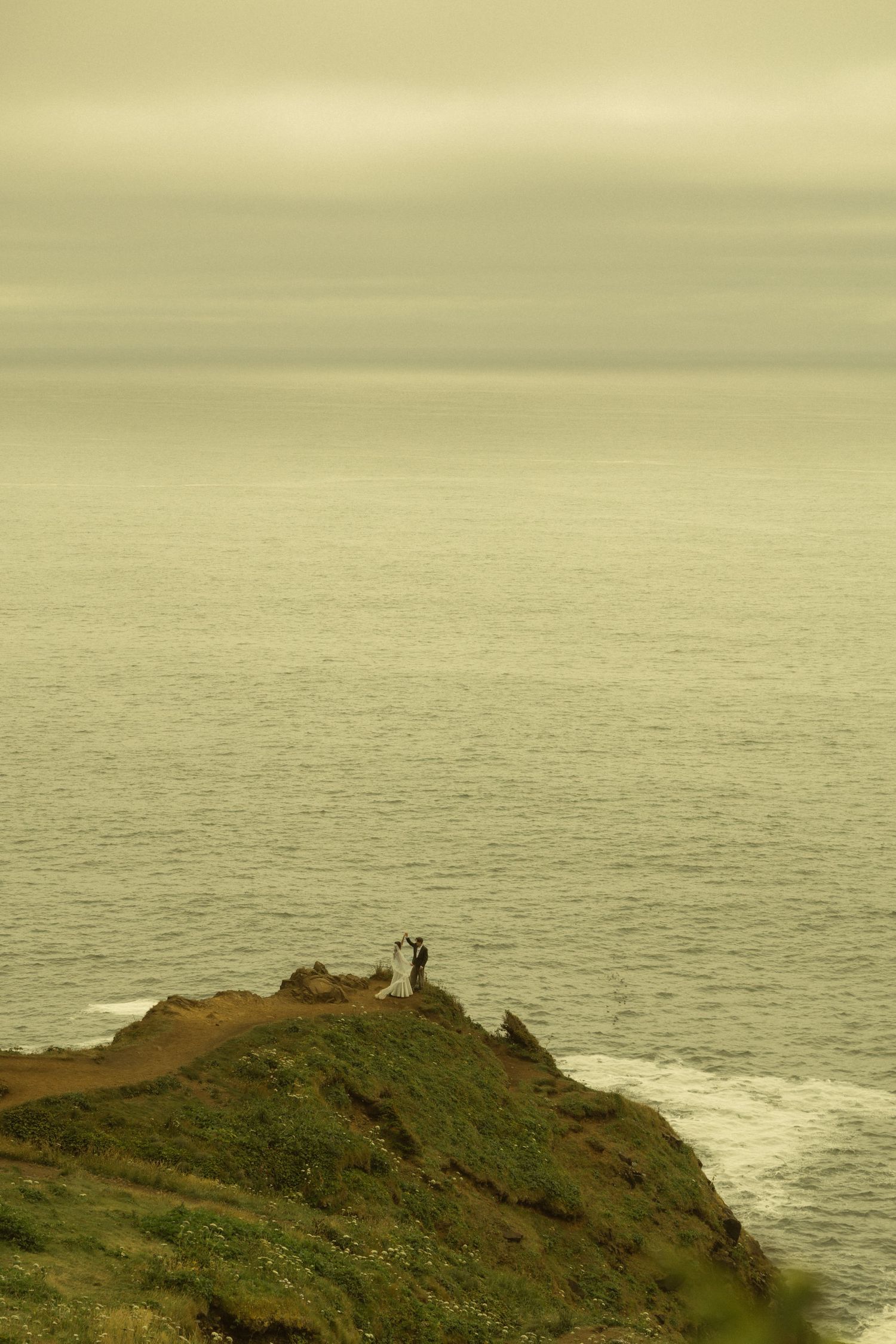 Couple standing on a coastal cliff overlooking a calm ocean under overcast skies.