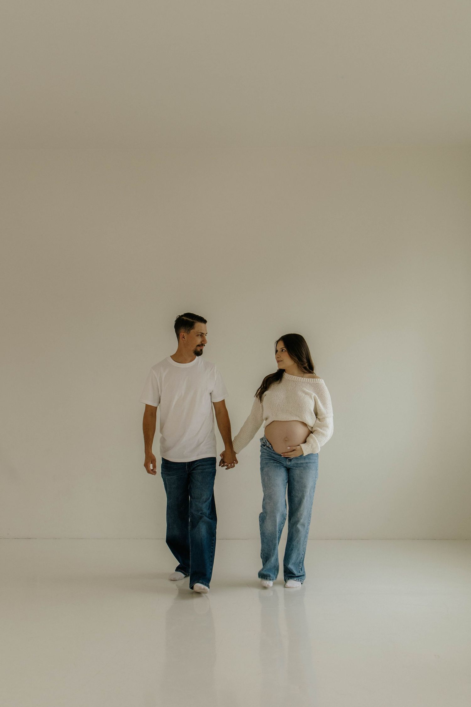 Minimalist maternity photo of couple walking hand in hand in white studio setting.