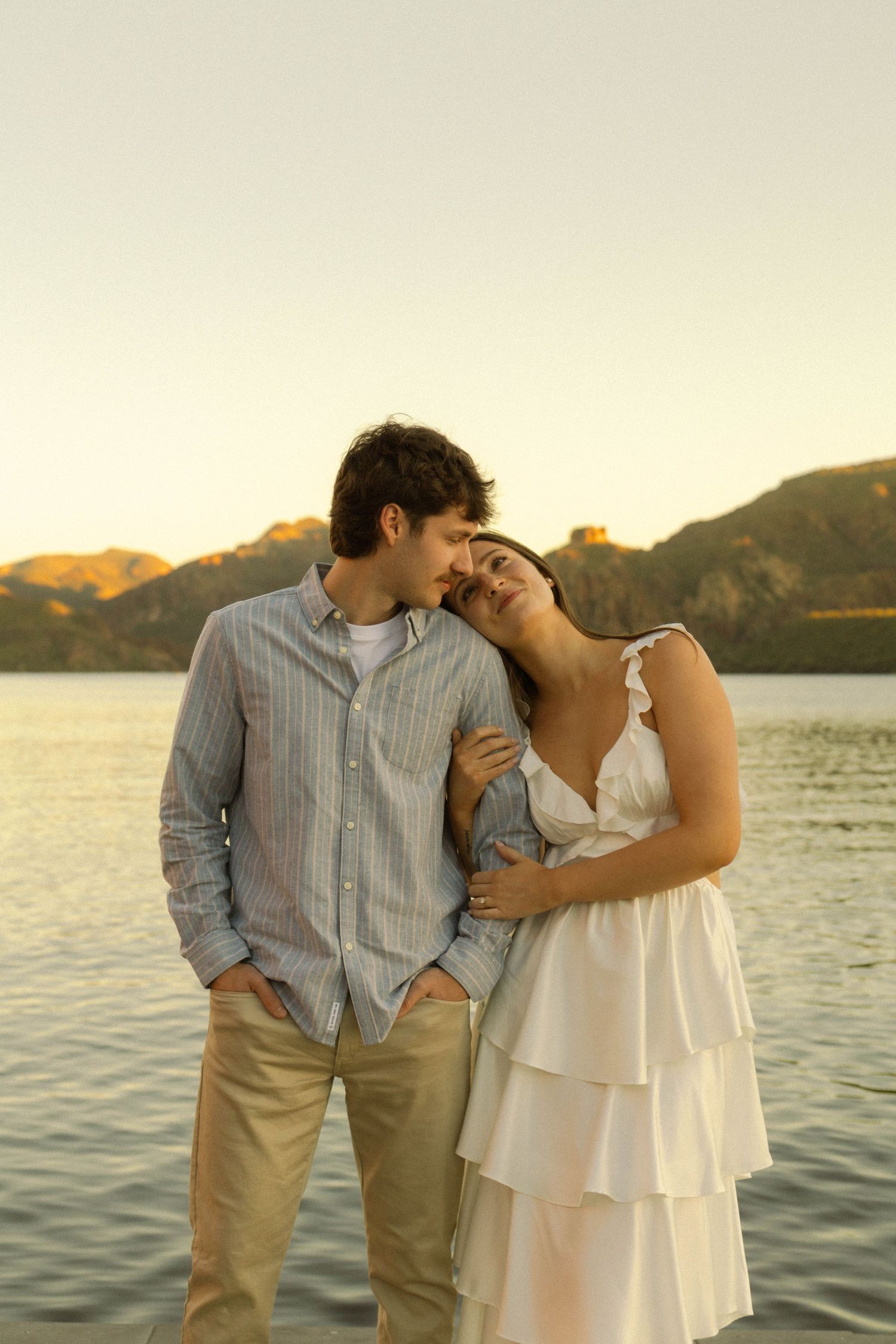 A romantic couple embraces by a scenic lake at sunset with mountains in the background.
