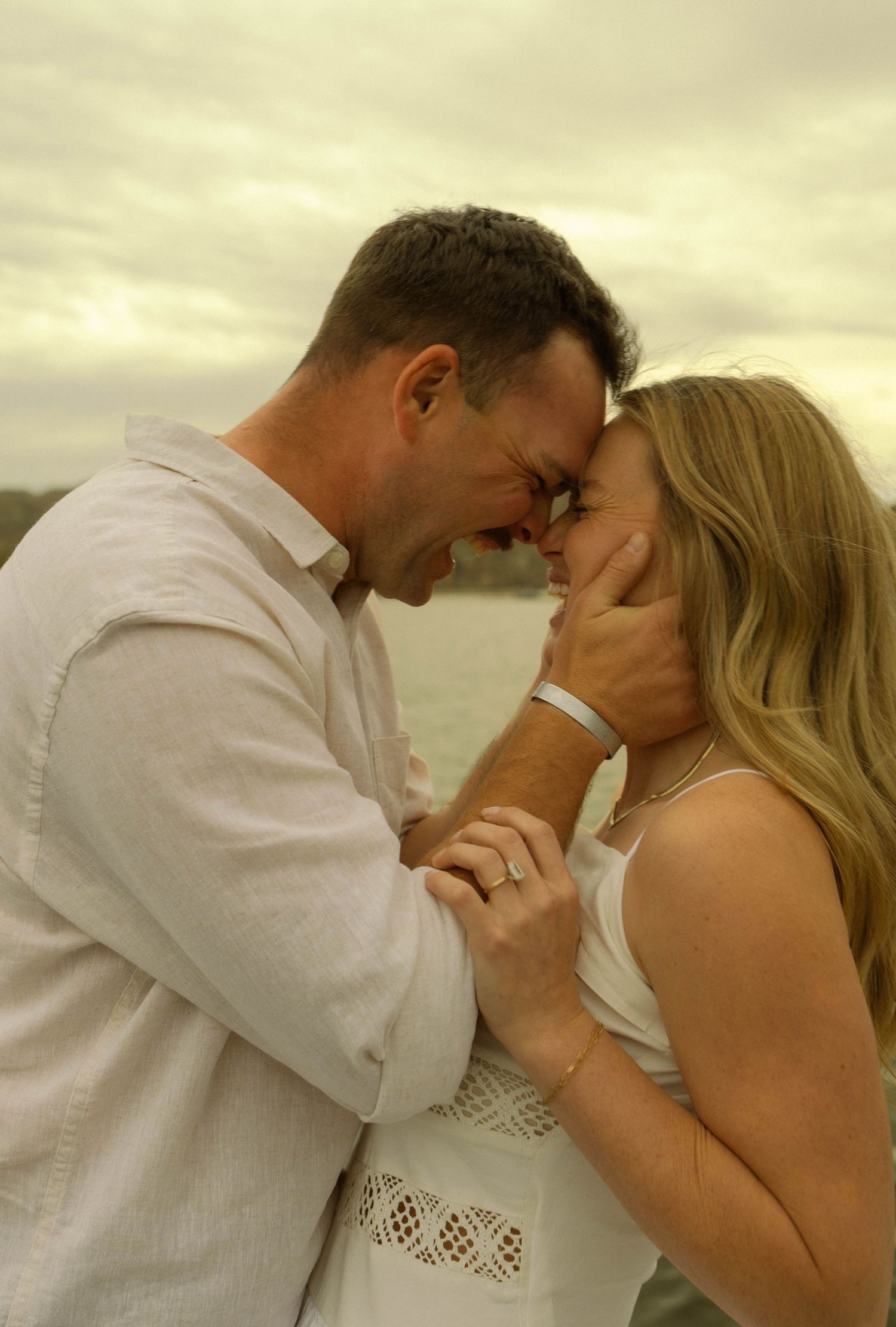 A romantic couple shares an intimate moment at sunset by the water wearing white clothing.