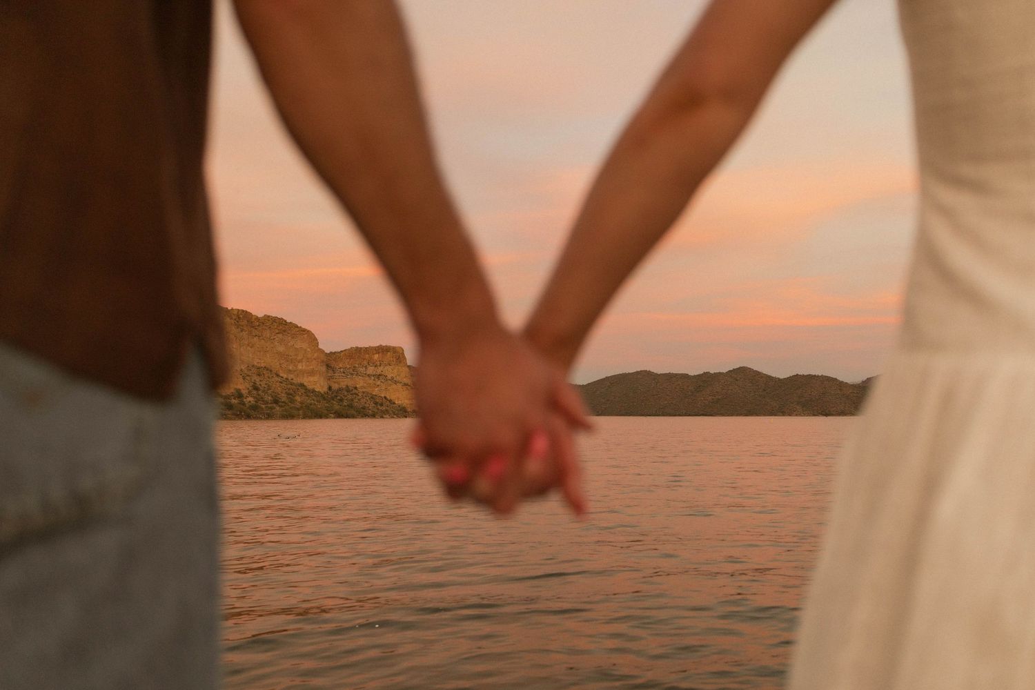 Couple holding hands while walking on beach at sunset with ocean and mountains in background.
