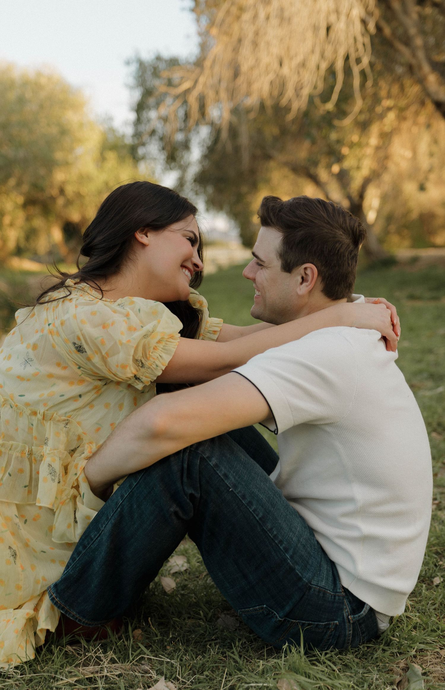 A playful couple laughs and embraces while sitting together on grass under golden sunlight.