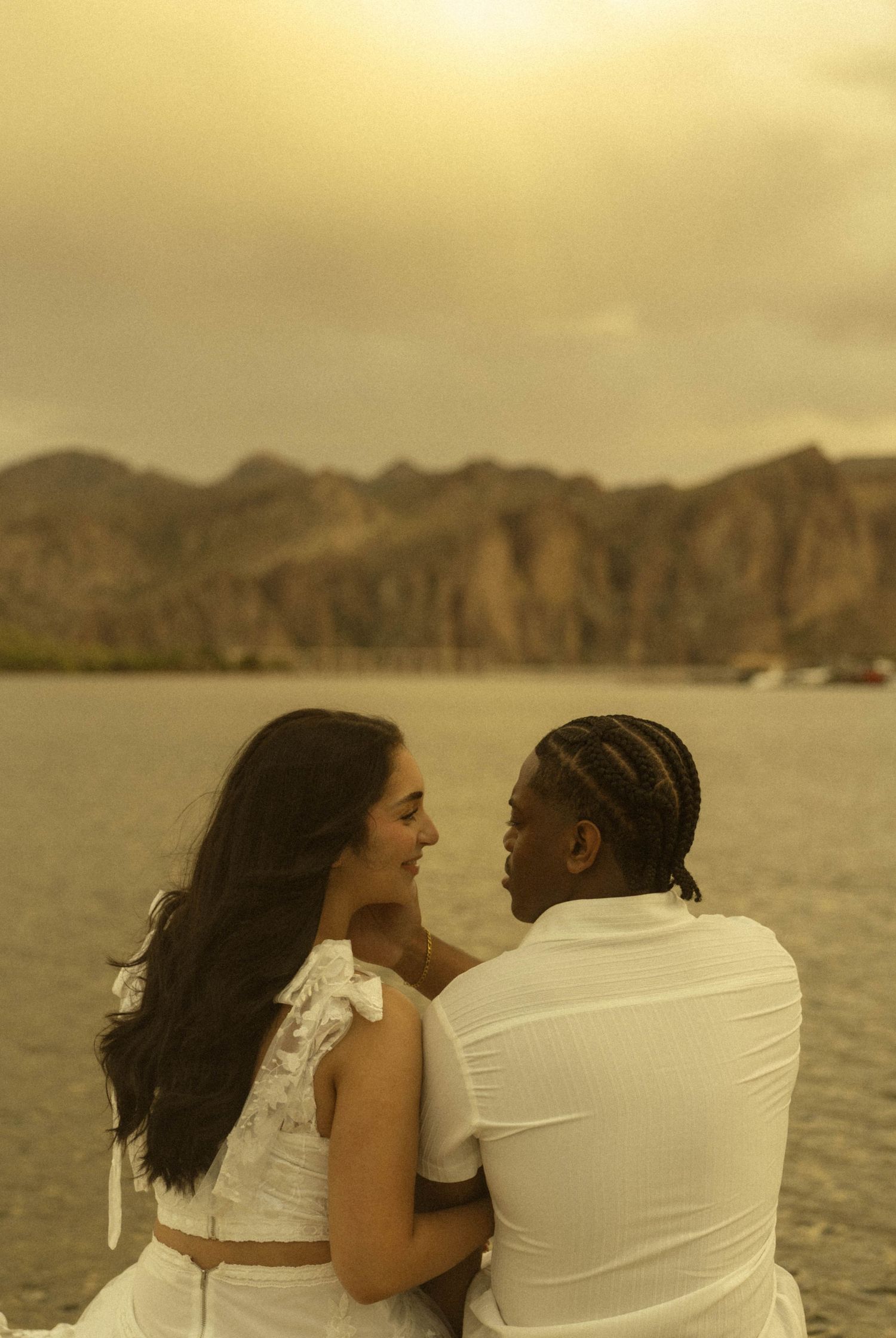 Romantic couple in white outfits sitting together gazing at each other by a mountain lake at sunset.