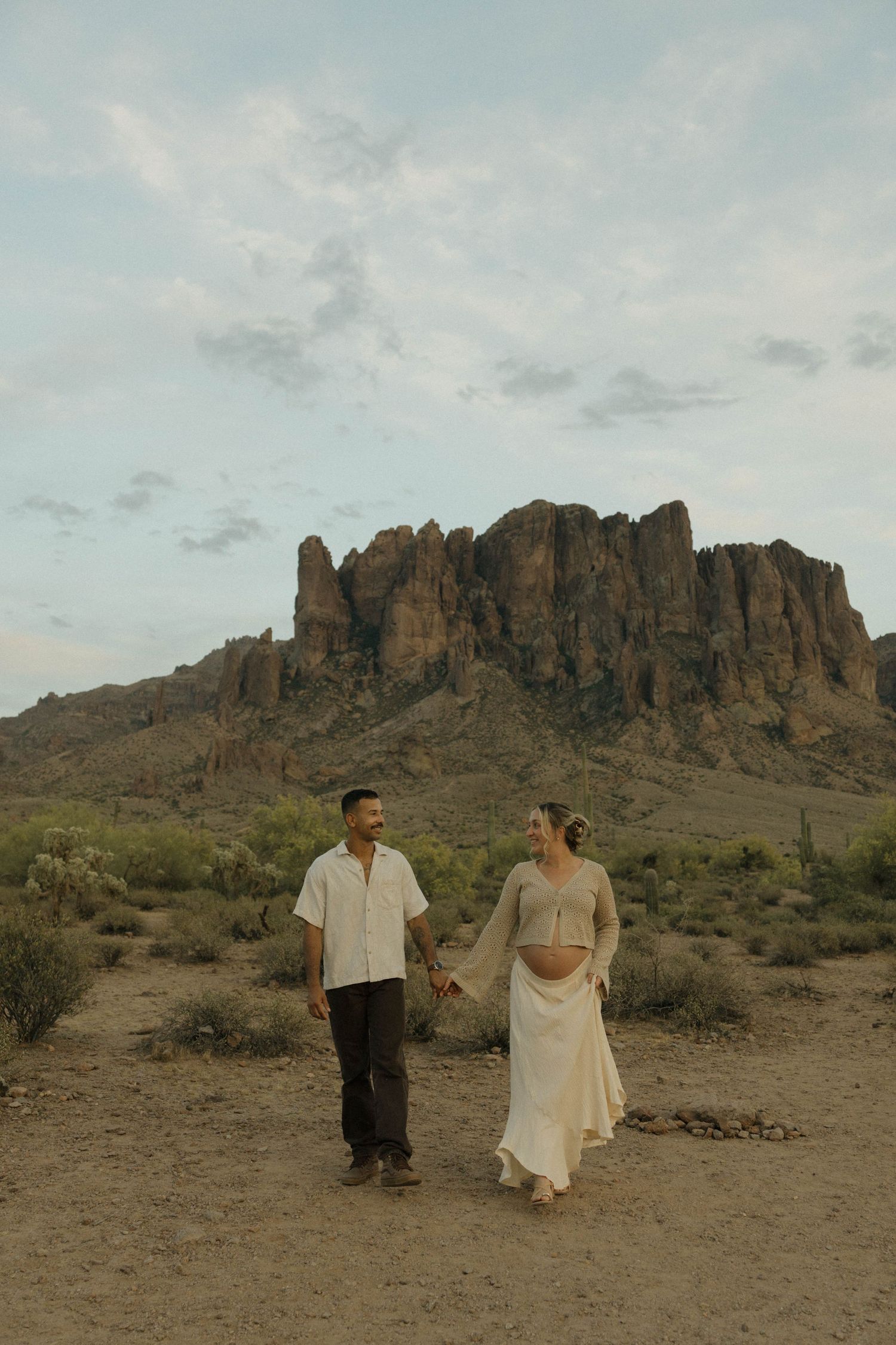Pregnant couple holding hands walking in Arizona desert with Superstition Mountains rocky peaks in background.