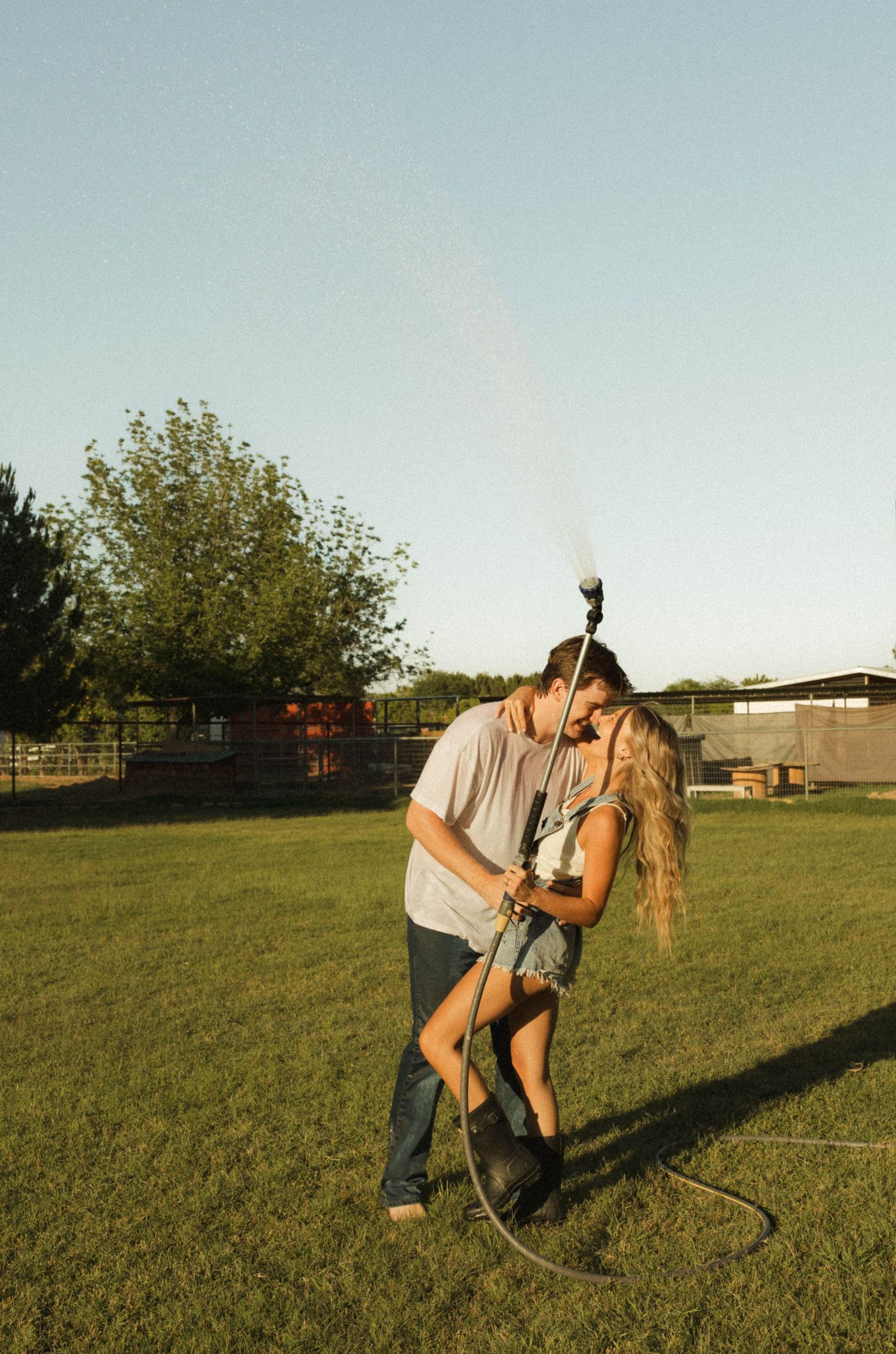 Couple sharing a romantic kiss while playing with a garden hose on a sunny green lawn.