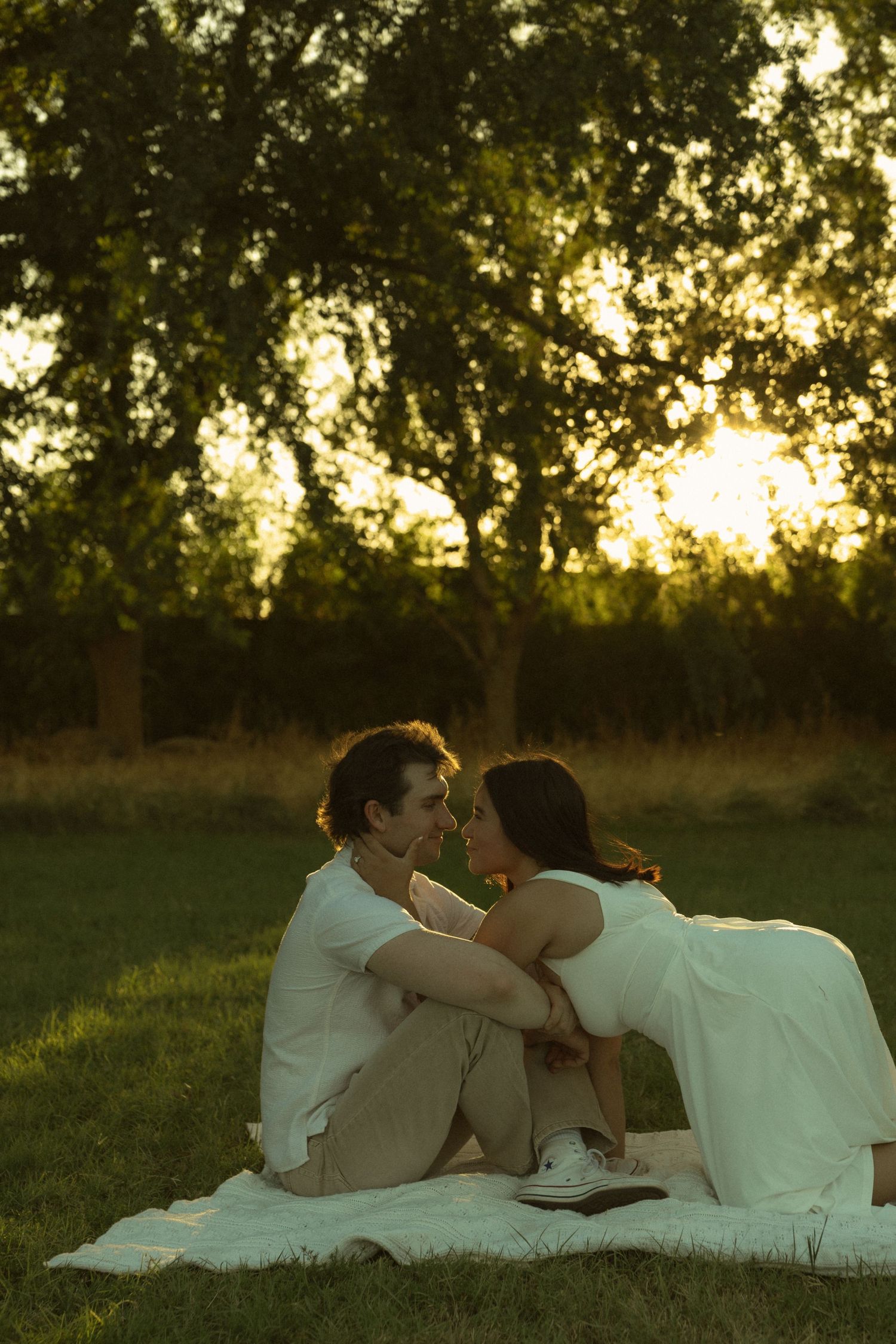 Romantic couple sitting close together on a blanket in a sunlit park at golden hour, nearly kissing.