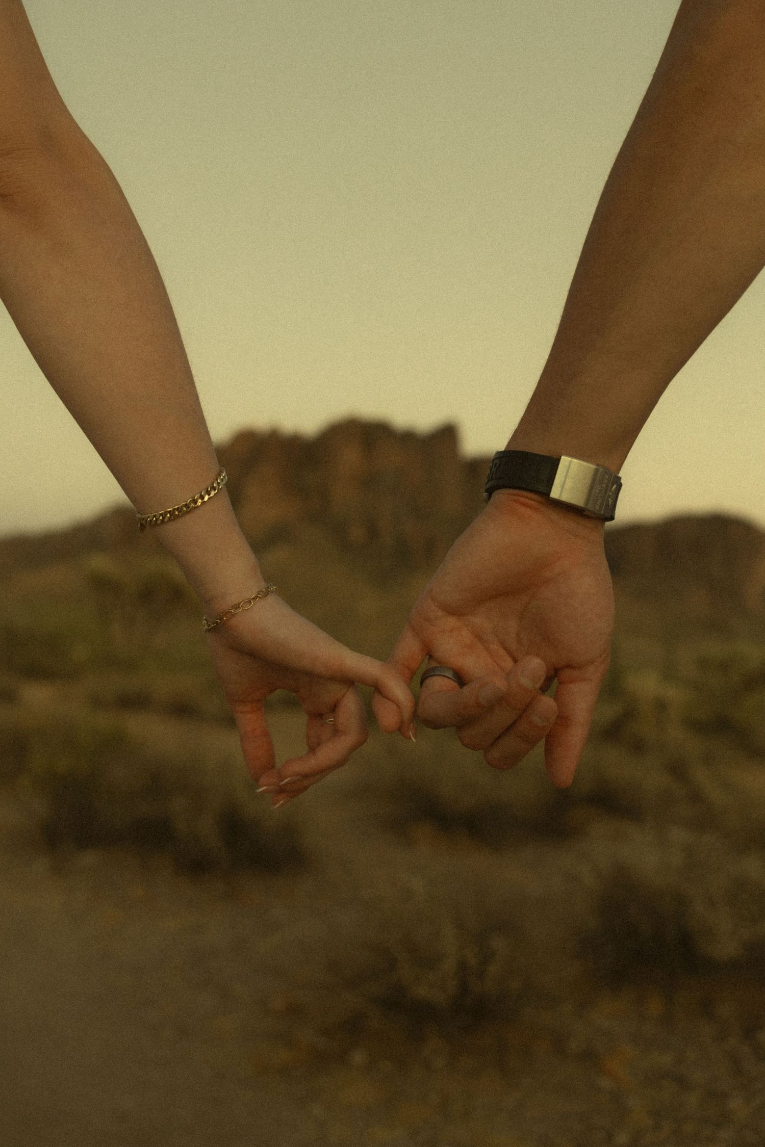Close-up of couple holding pinky fingers with desert mountain scenery and warm golden light in background.