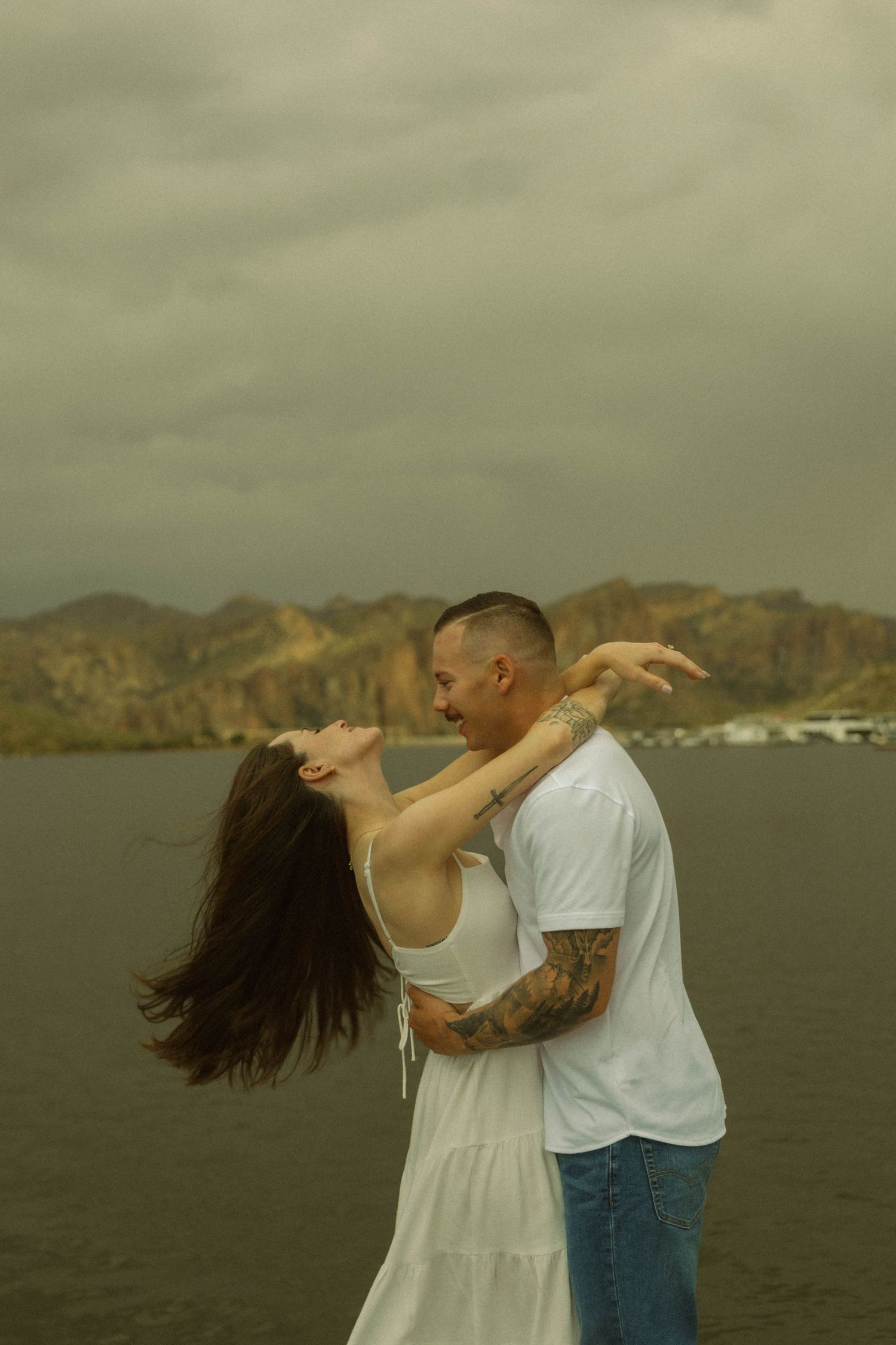 A couple dances together against a scenic mountain and water backdrop in romantic sunset lighting.