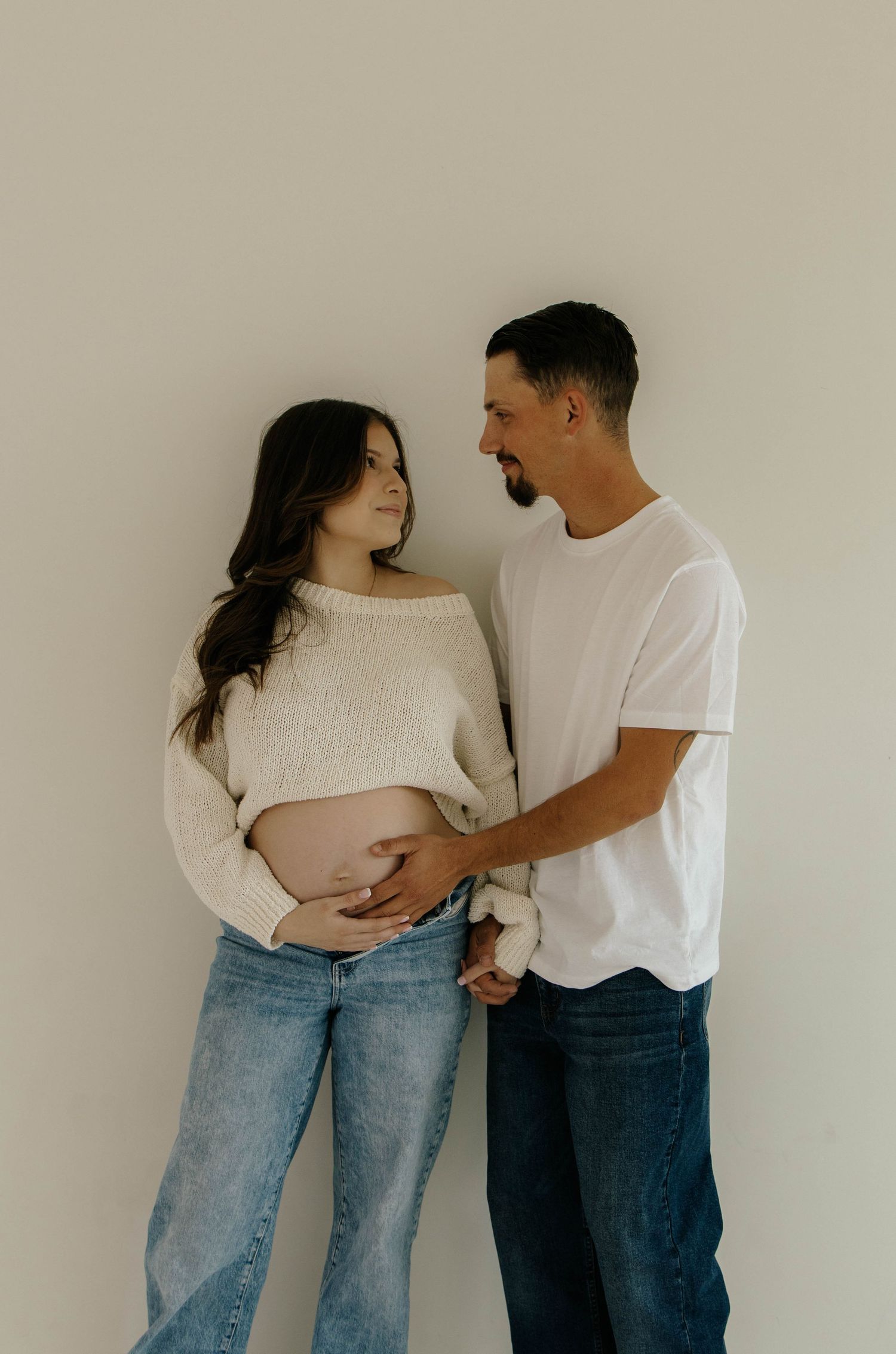 Expecting couple in casual outfits embracing and showing pregnancy in minimalist studio setting.