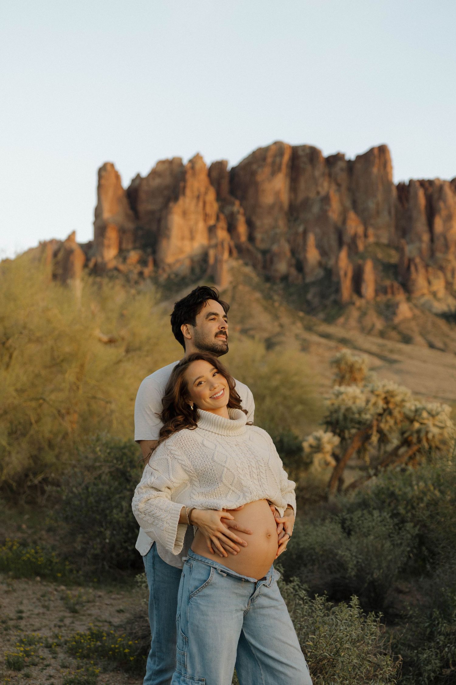 A couple embraces against a backdrop of dramatic desert cliffs and mountains during golden hour.