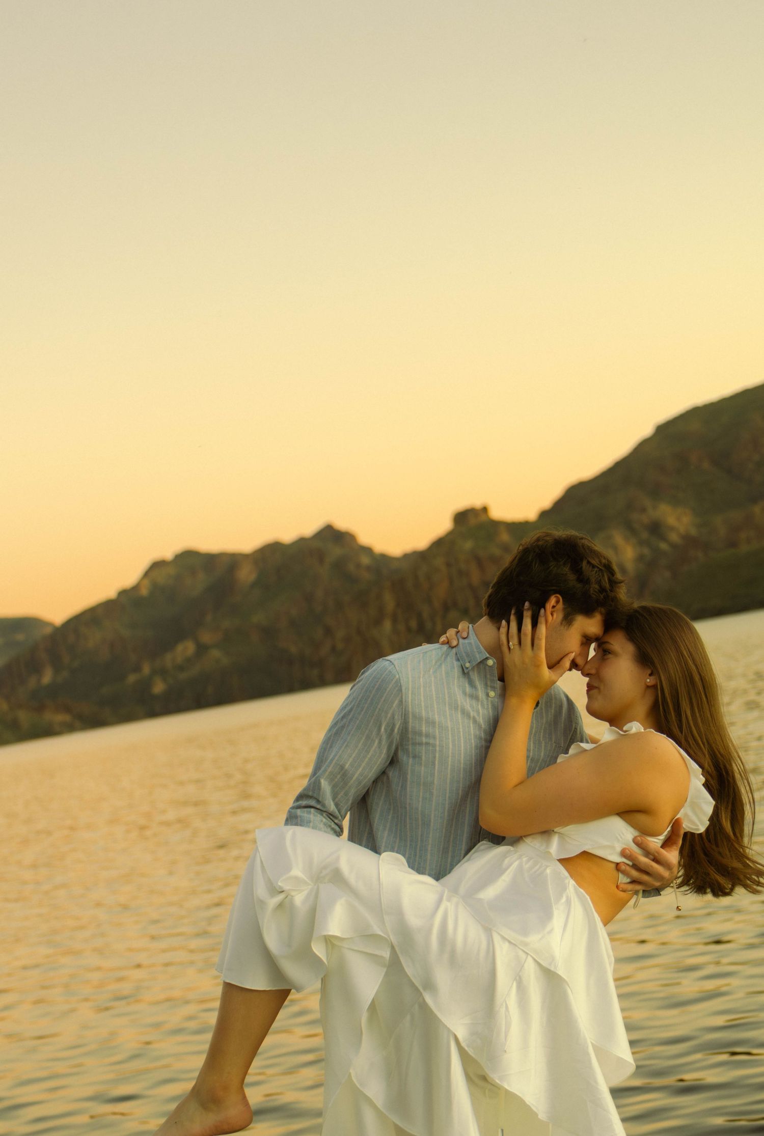 Two people in white and blue clothing share a romantic embrace against a sunset lake backdrop.