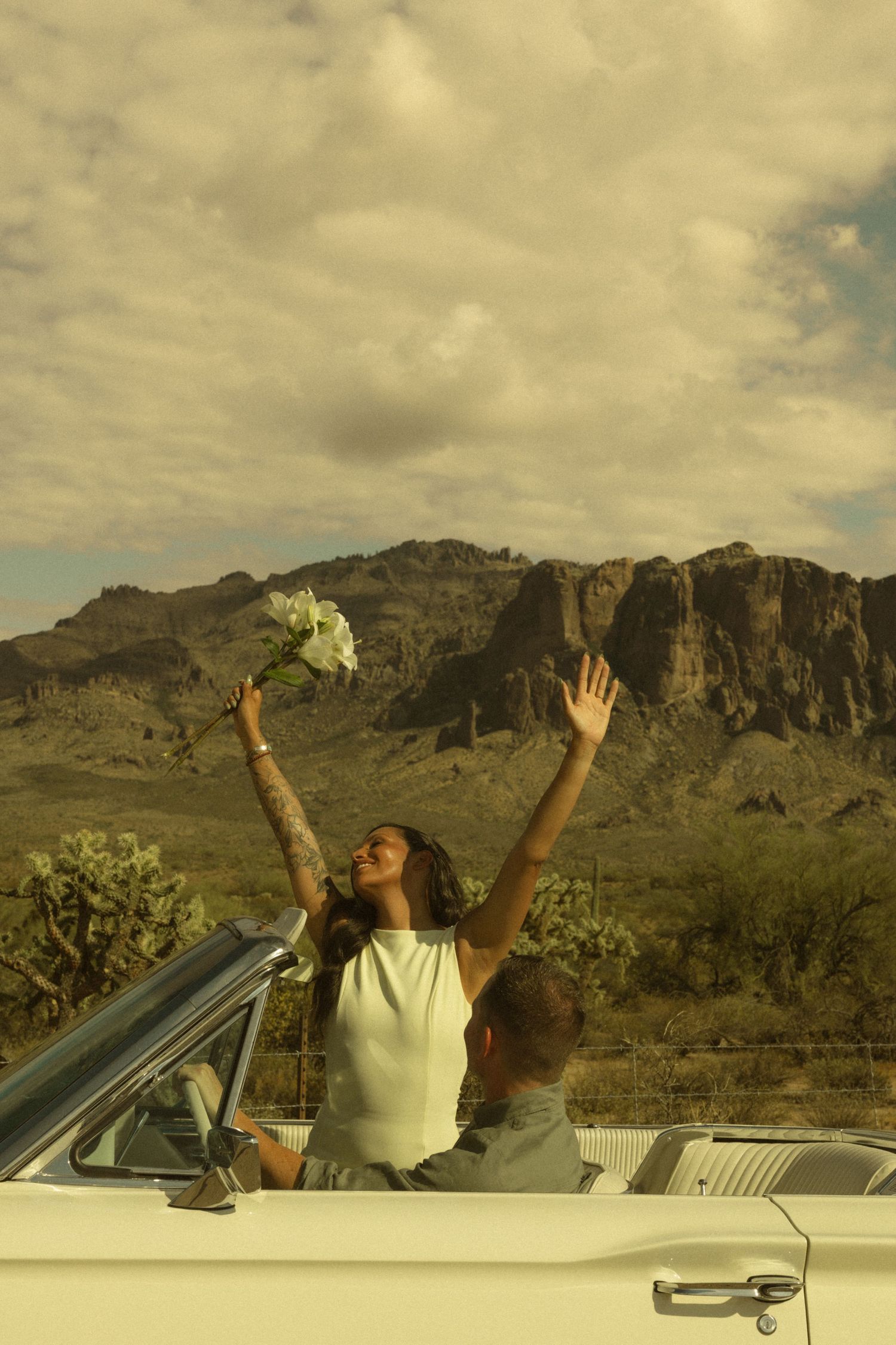 Joyful woman raising bouquet in white convertible with desert mountain scenery behind.