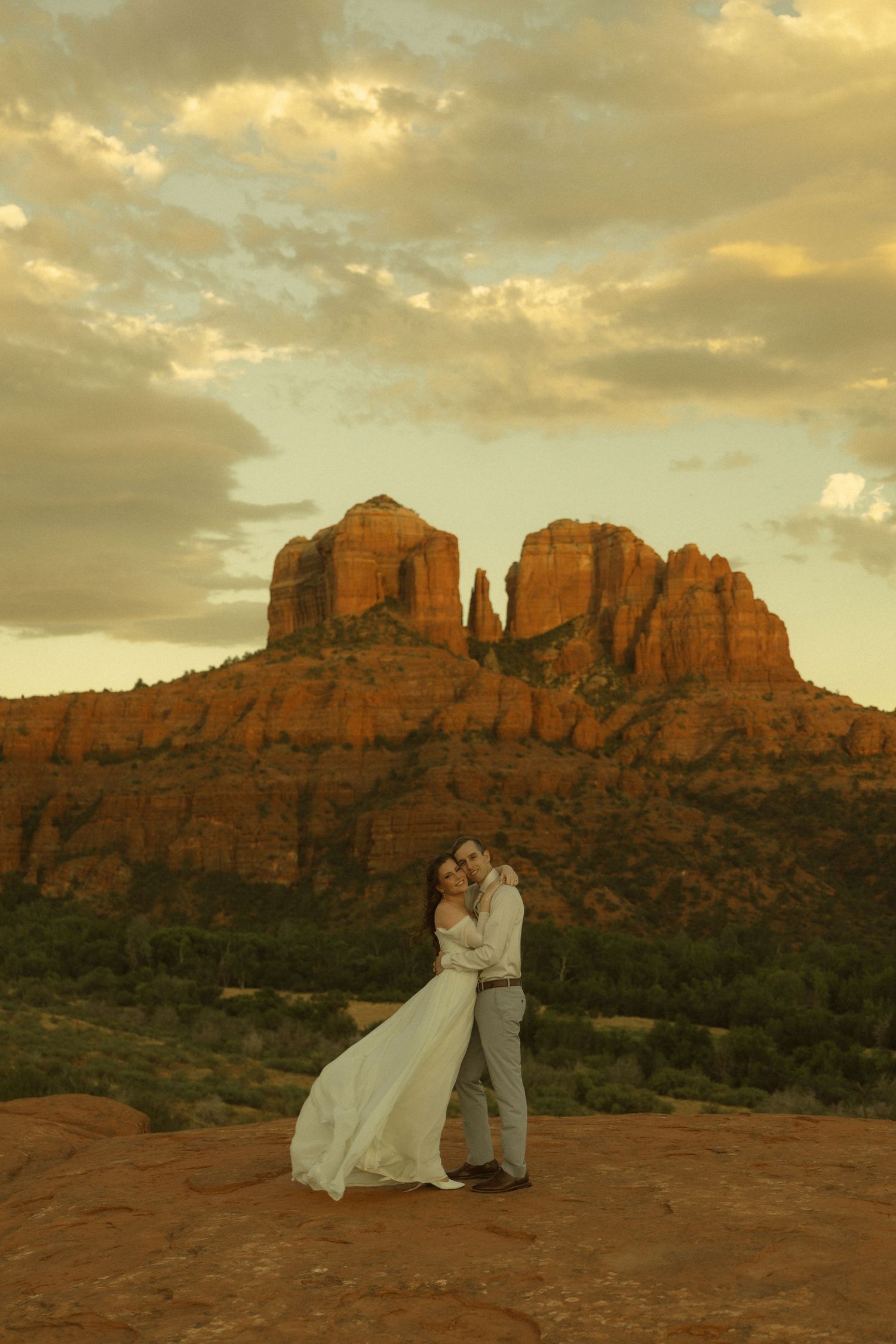 Couple embraces in wedding attire at sunset with Cathedral Rock formation in Sedona, Arizona glowing red in background.