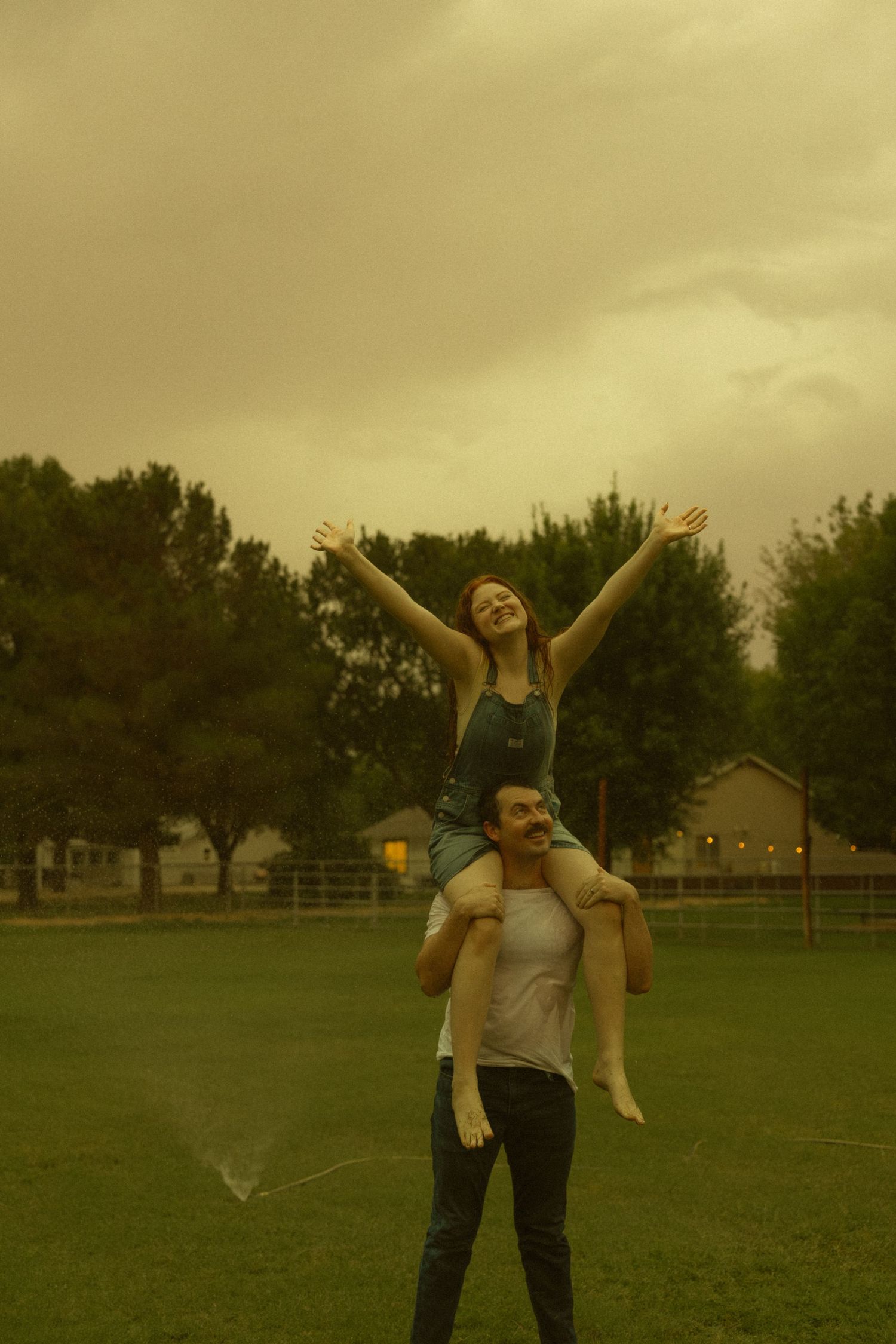 A woman with arms raised sits on a man's shoulders in a park under a dramatic stormy yellow sky.
