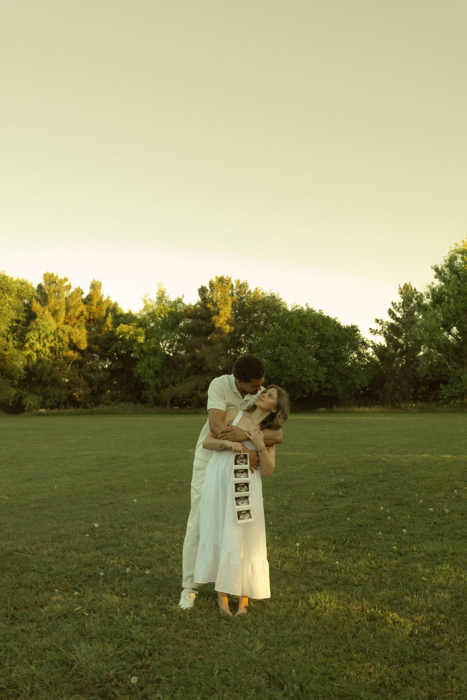 Couple embracing in a sunlit park, woman in white dress holding ultrasound photos, golden hour maternity announcement.
