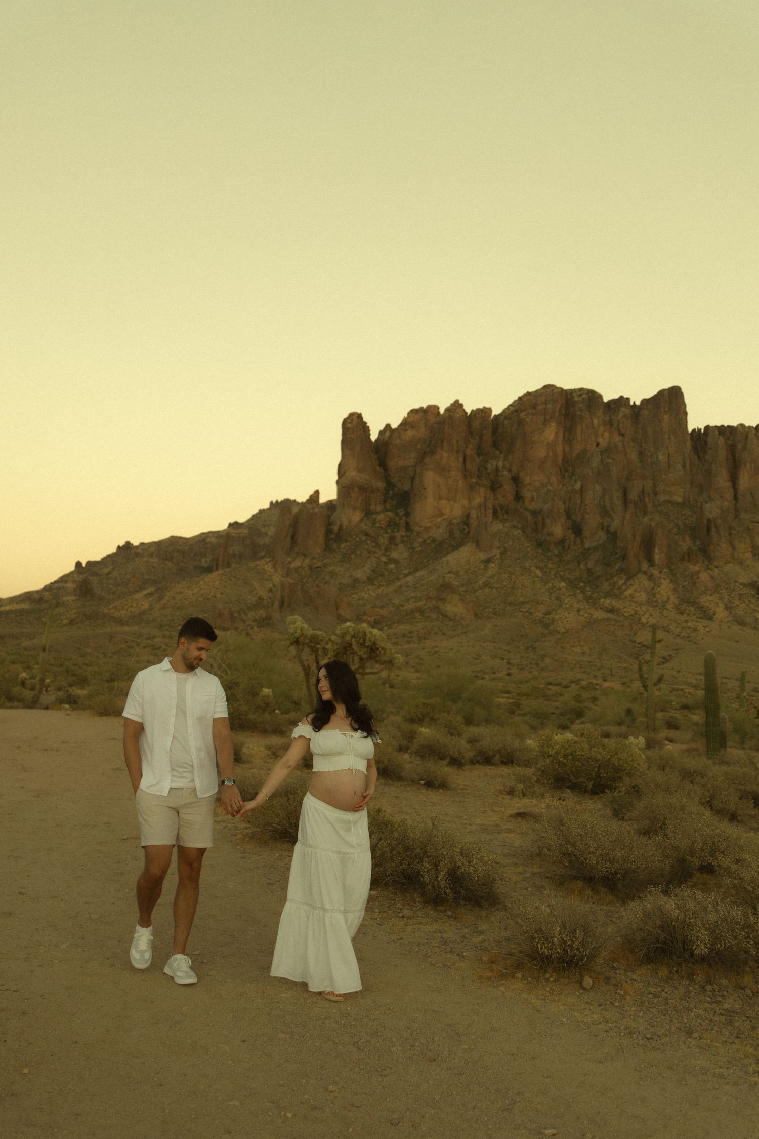 Pregnant couple holding hands in white outfits at desert sunset with Superstition Mountains in background.