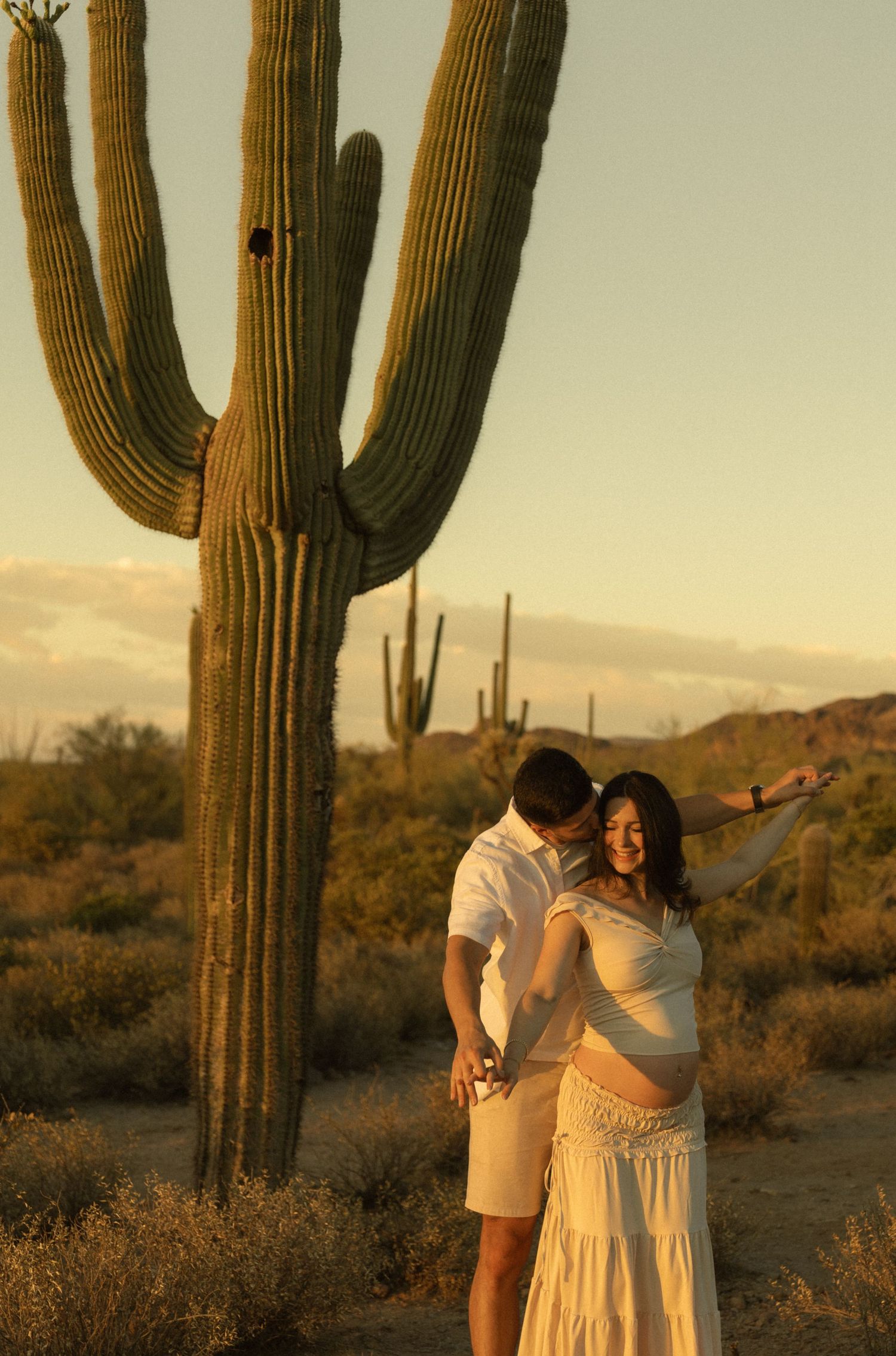 Couple embracing and laughing during maternity photoshoot in Sonoran Desert at sunset near saguaro.