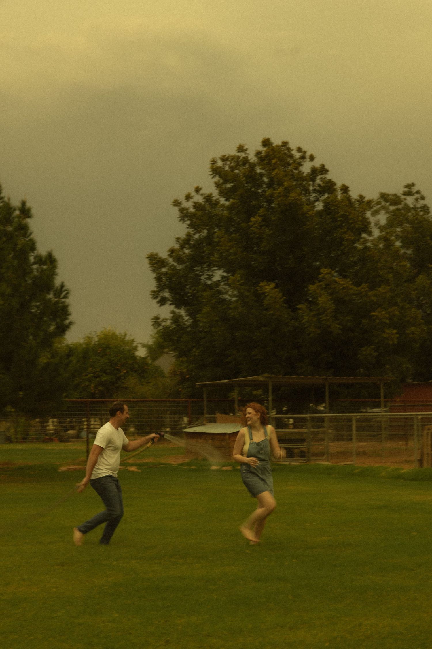 Couple playing and running barefoot on green grass in a park with trees under a dramatic cloudy sky.