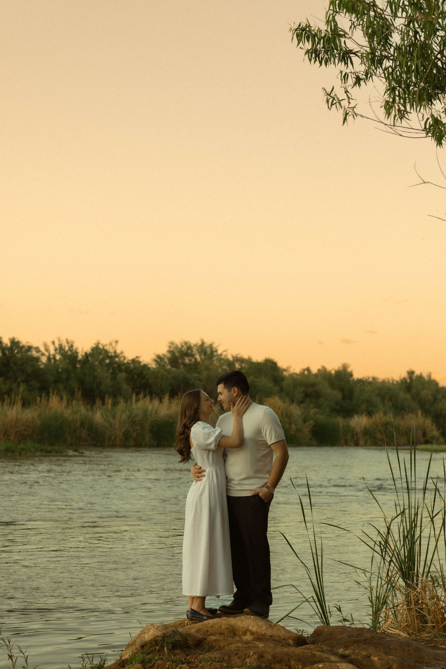 Couple tenderly facing each other on rocks by a tranquil river at sunset, woman in white dress caressing mans face.