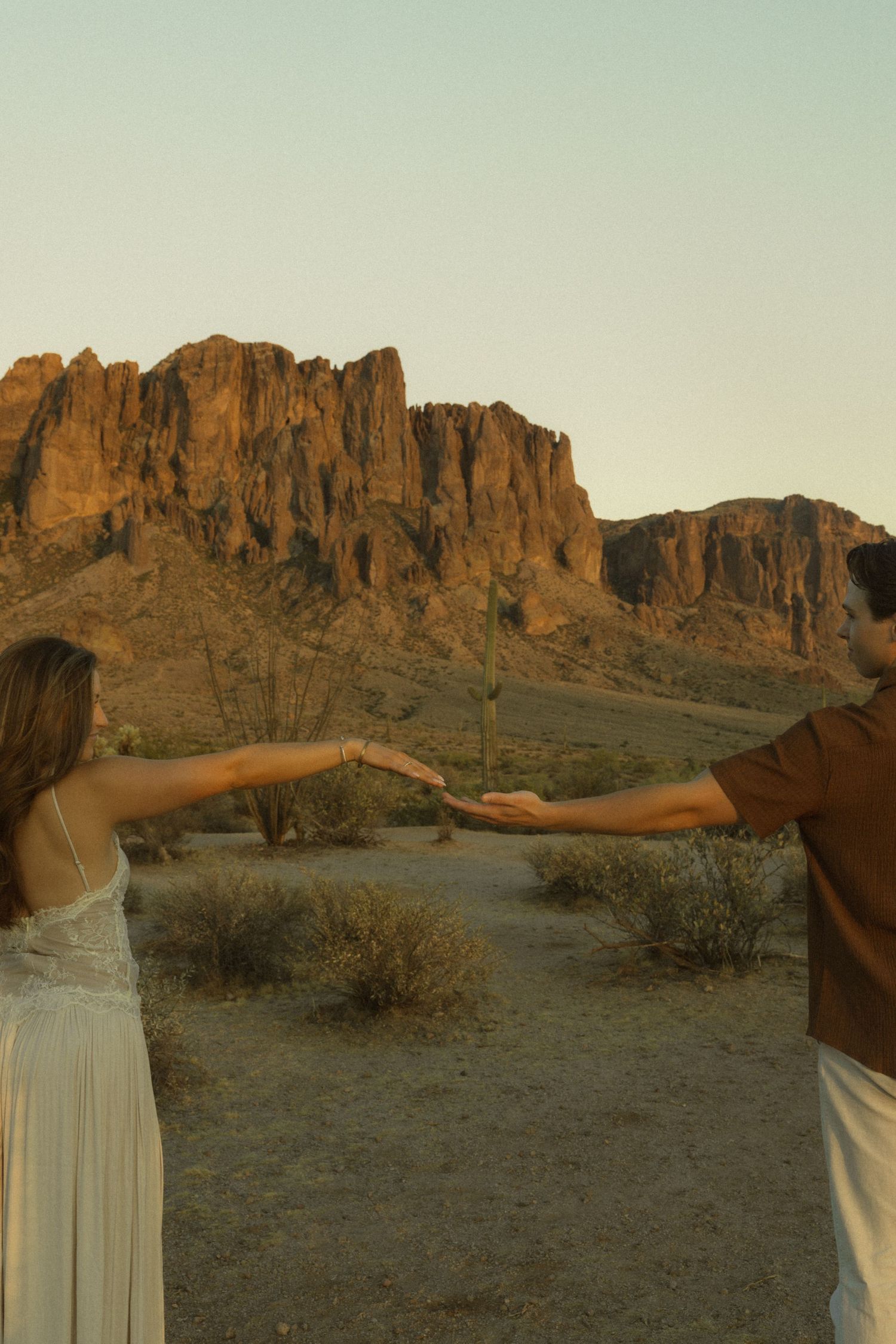 Couple reaching toward each other in desert landscape with dramatic red rock mountain formation at golden hour.