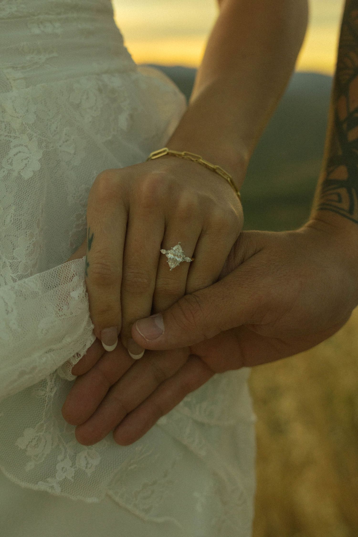 Close-up of engagement ring on bride's hand with groom holding her hand at sunset.