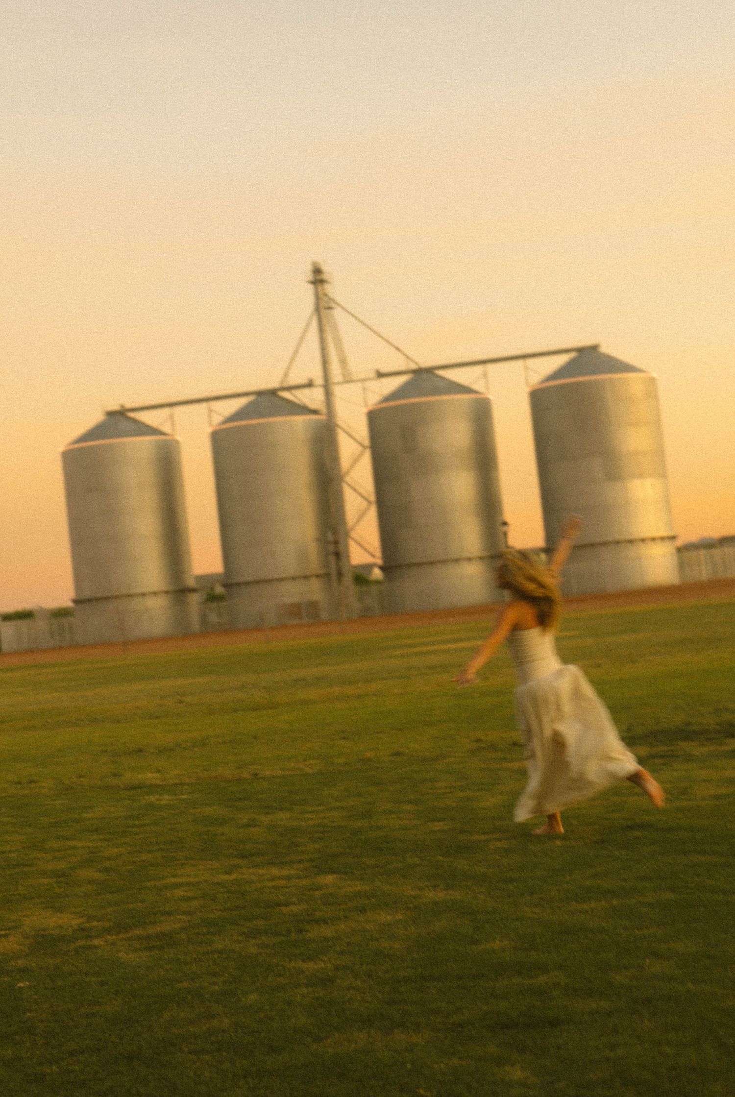 A woman in a white dress runs across a green field with large silver grain silos in the background at sunset.