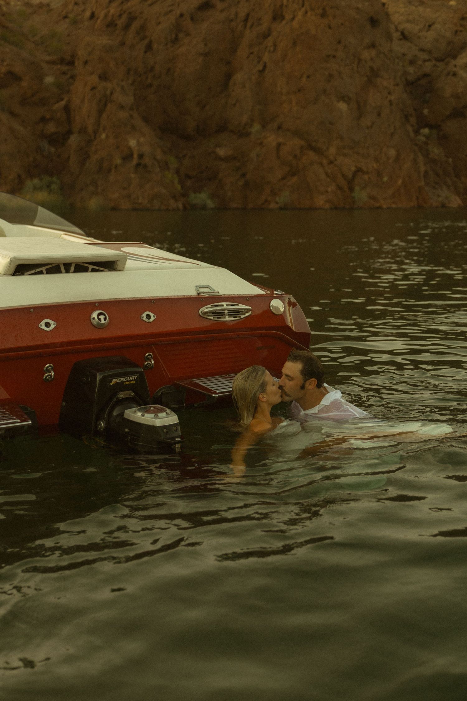 Romantic couple kissing in the water beside a red and white speedboat in a canyon lake.