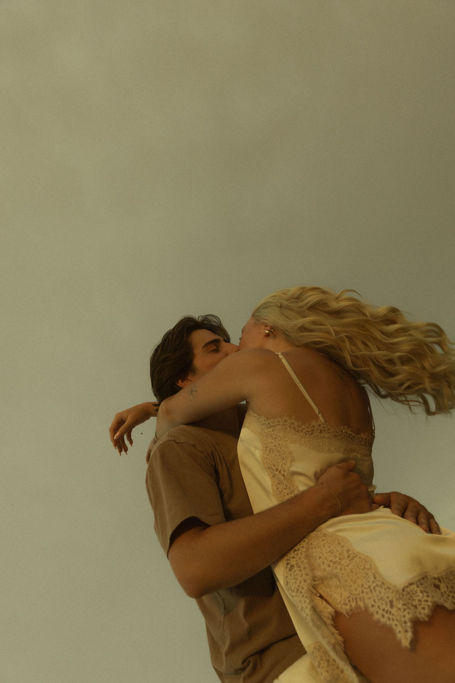 Romantic couple kissing while man lifts woman in flowing dress against a soft neutral background.