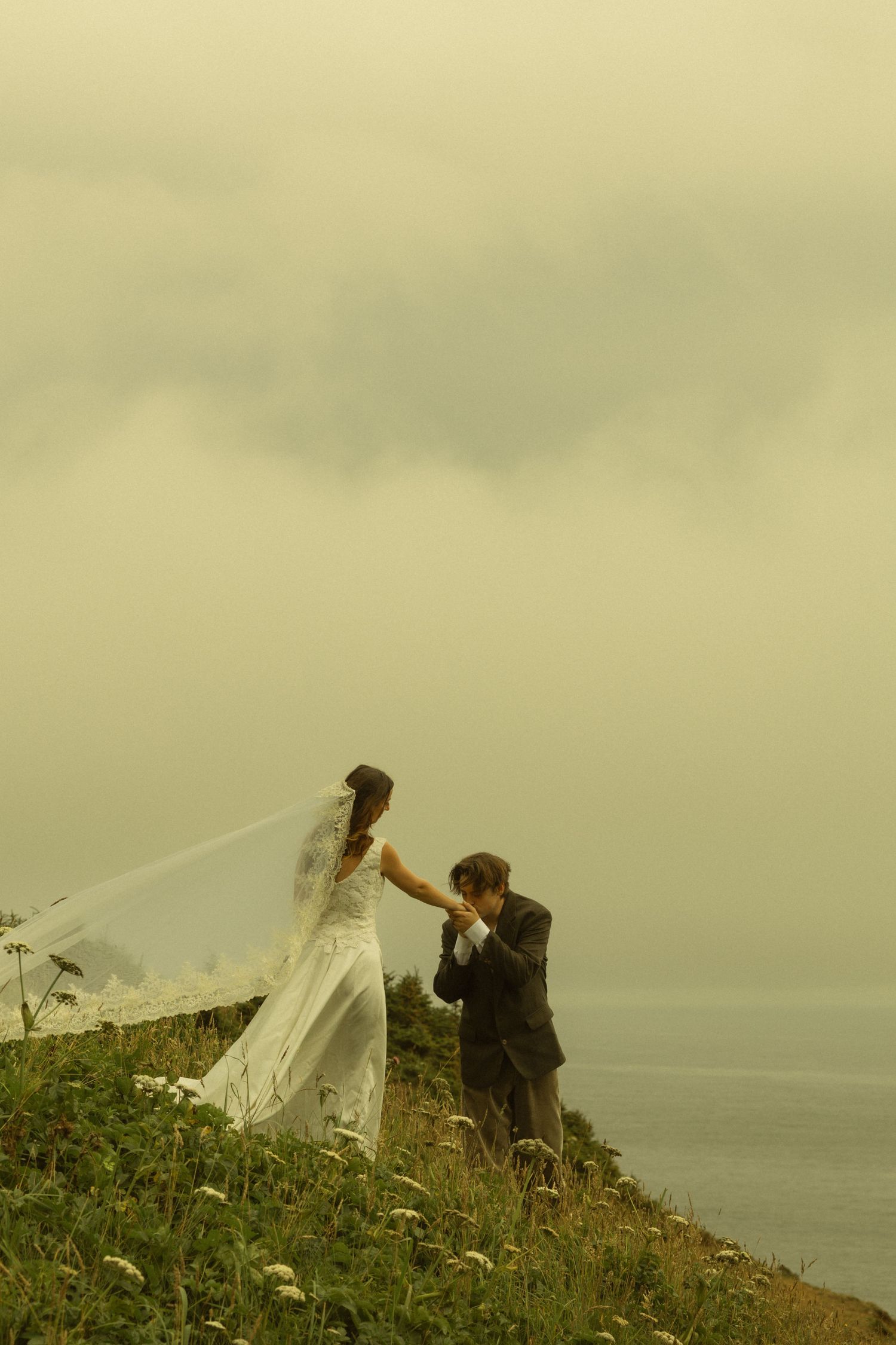 Groom kissing bride's hand on a misty cliff overlooking the sea during an outdoor wedding ceremony.