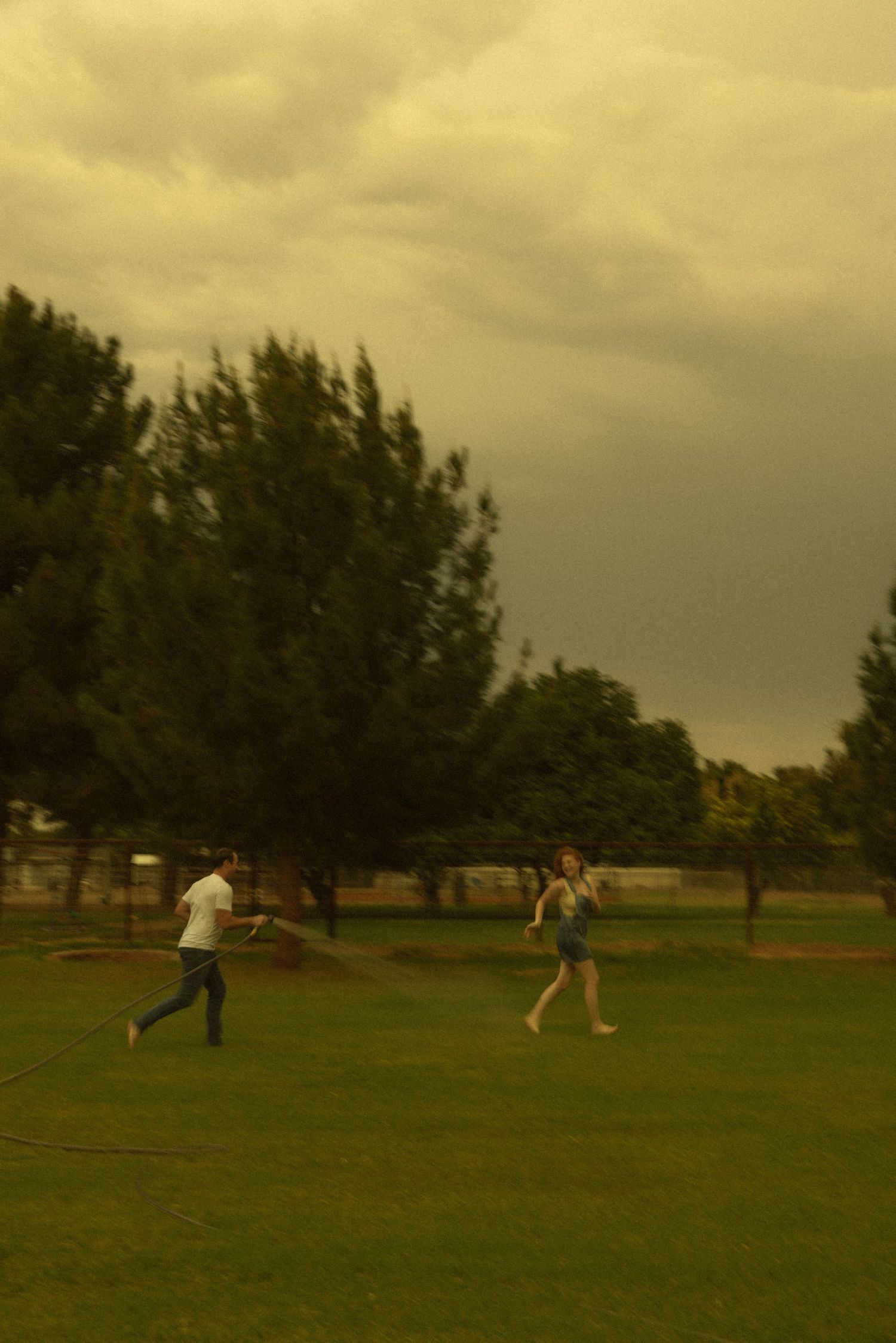Two people playing in a lush green park under dramatic stormy skies with tall trees in background.