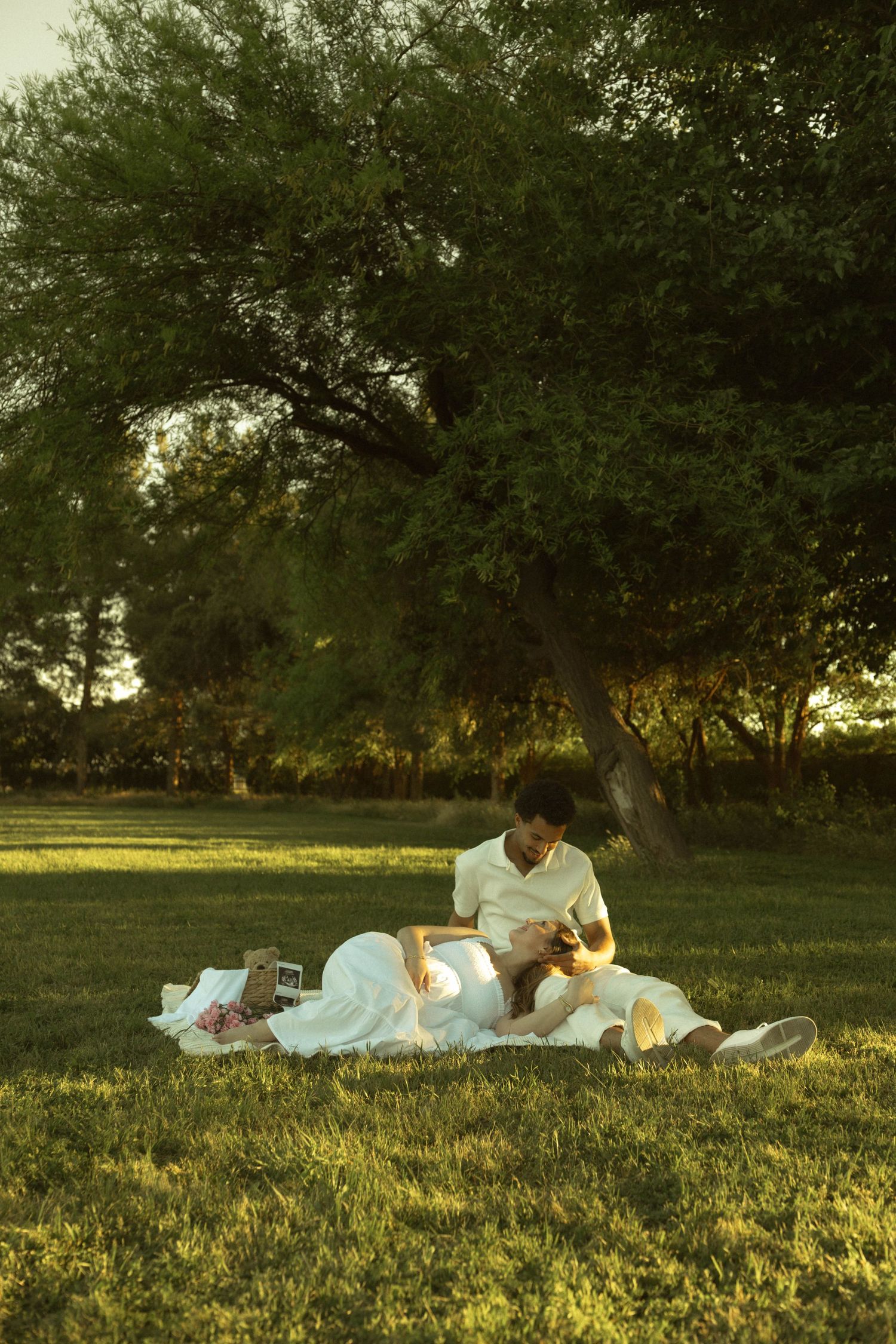 Couple in white outfits enjoying a romantic picnic on a blanket in a lush green park at golden hour.