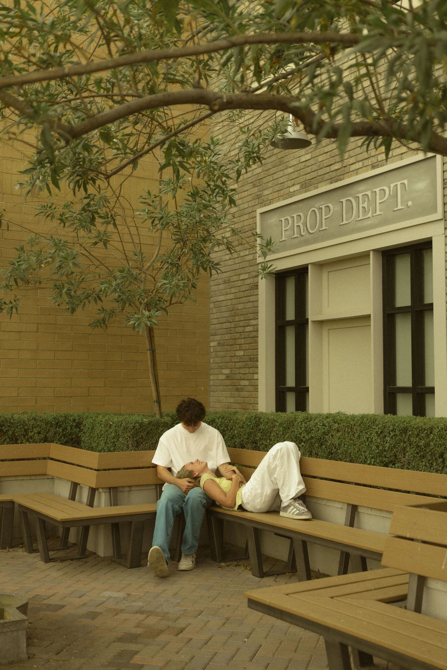 Couple relaxing on outdoor bench near Prop Dept building under tree.